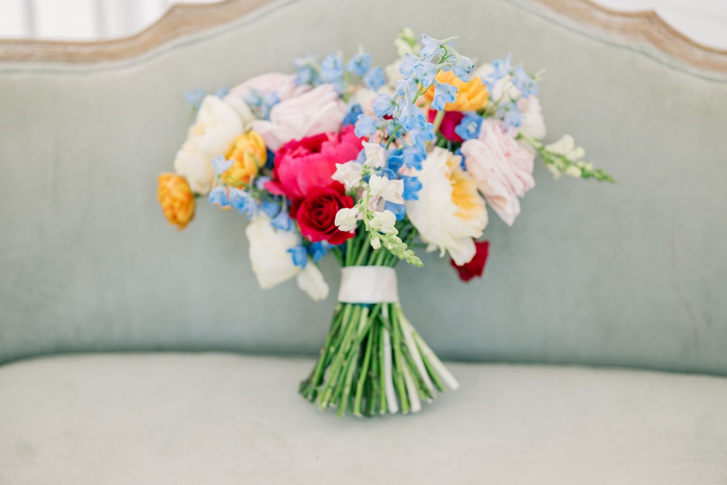 Colorful wedding bouquet with red, pink, yellow, and blue flowers resting on a vintage mint green sofa.