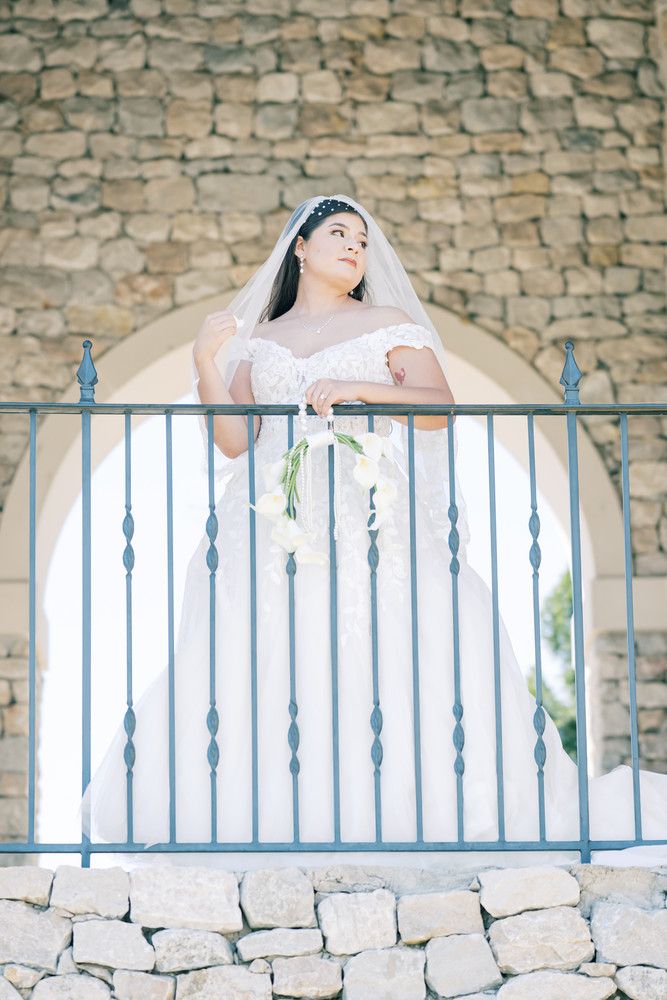 A bride in a white gown poses against a rustic stone wall with a metal railing at sunset.