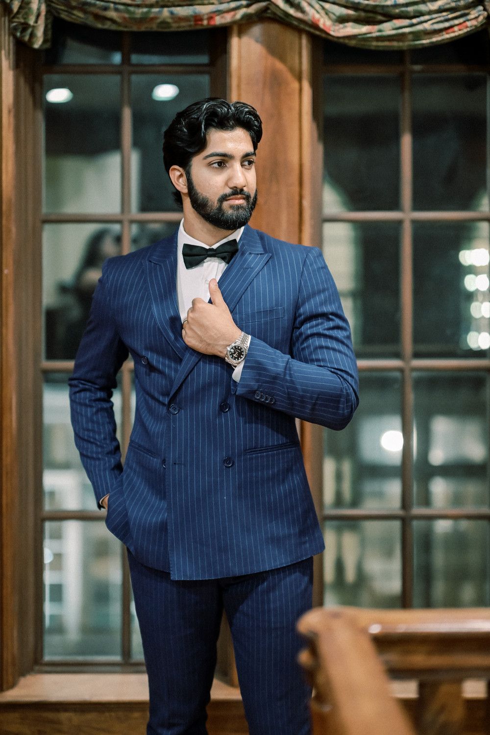 Man in pinstriped navy suit adjusts bowtie while posing in elegant interior.