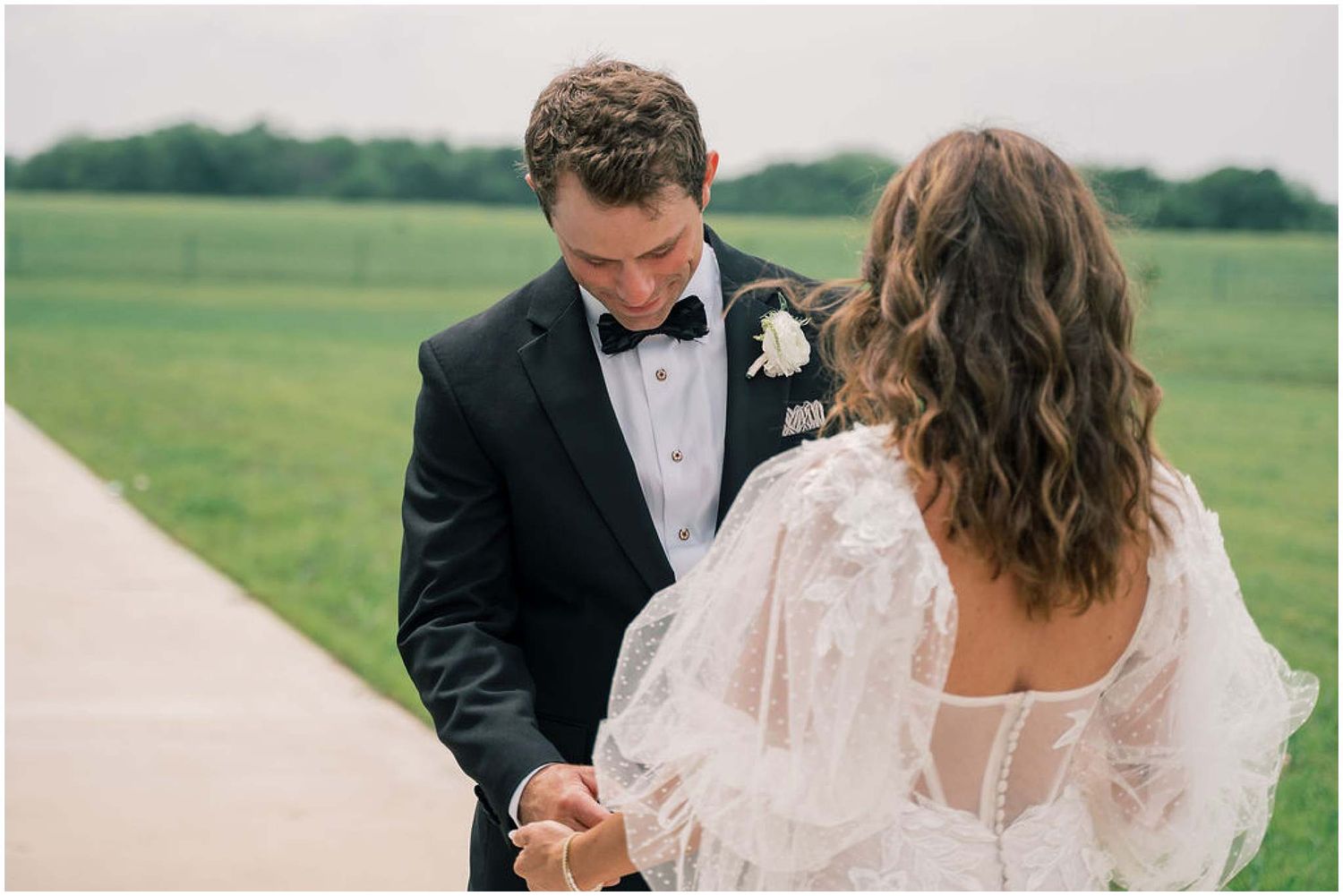 A couple shares an intimate moment on their wedding day in a grassy field, with the bride wearing a flowing lace gown.