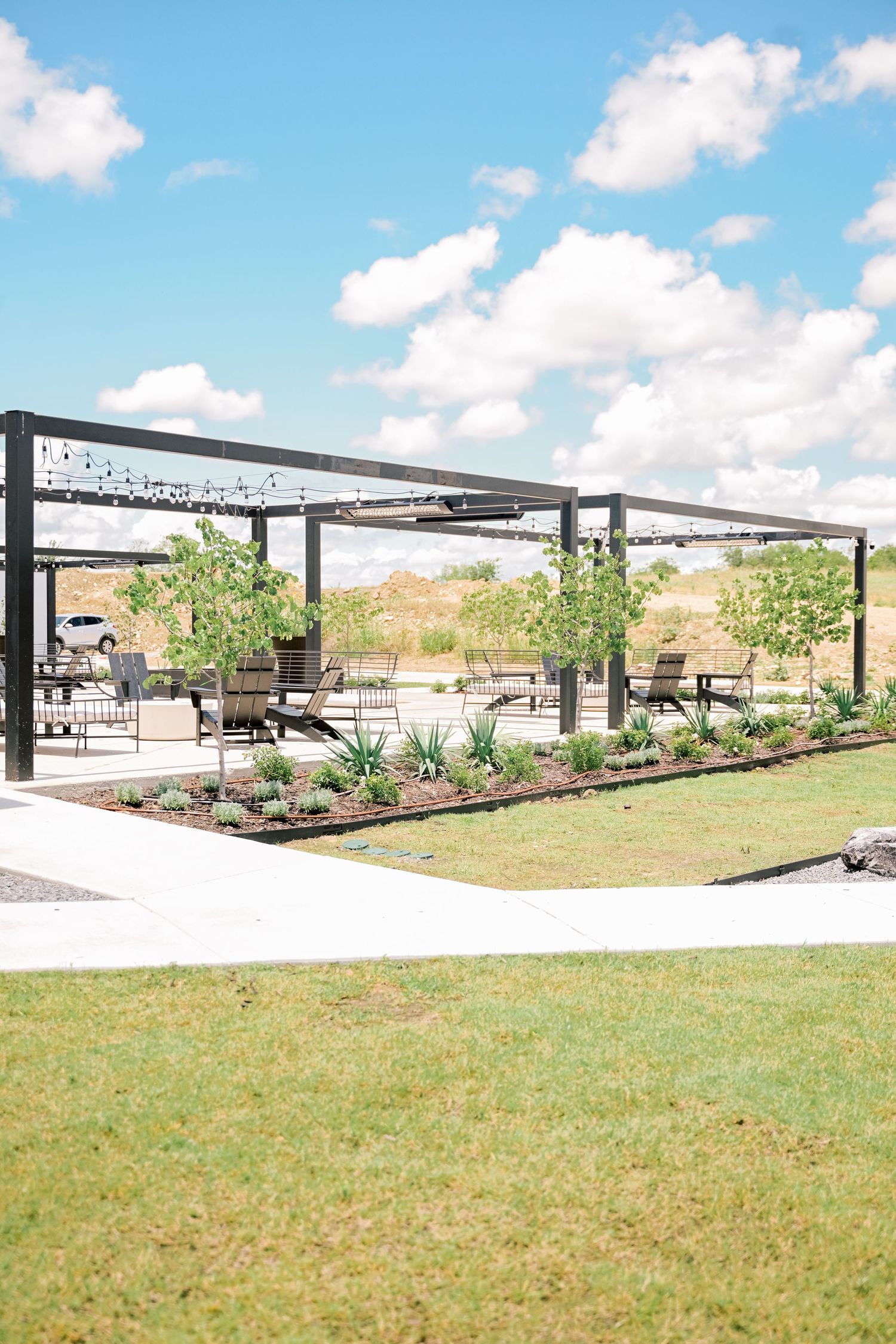 Modern outdoor wedding venue pergola with string lights, lush landscaping, and seating under blue sky.