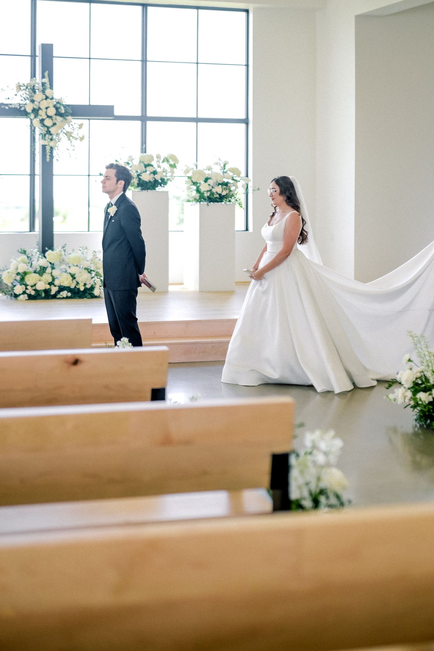 Bride in flowing white ball gown approaches groom for first look in bright modern chapel.