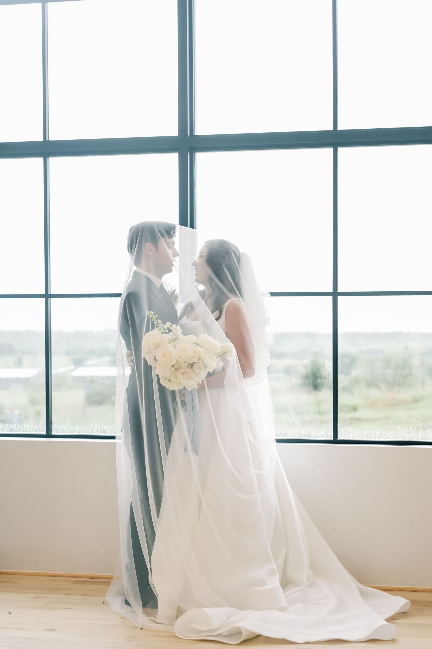 Bride and groom share an intimate moment under a flowing veil before large windows on their wedding day.