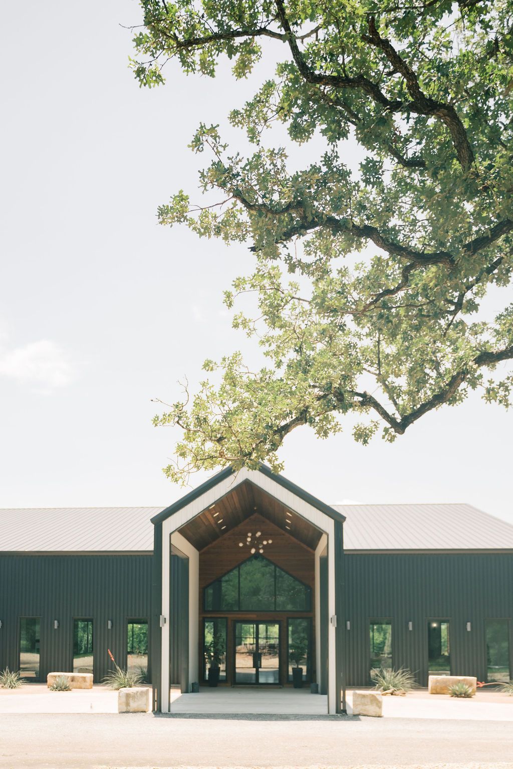 Dark green modern barn wedding venue with vaulted entrance, glass doors, and large oak tree overhead.