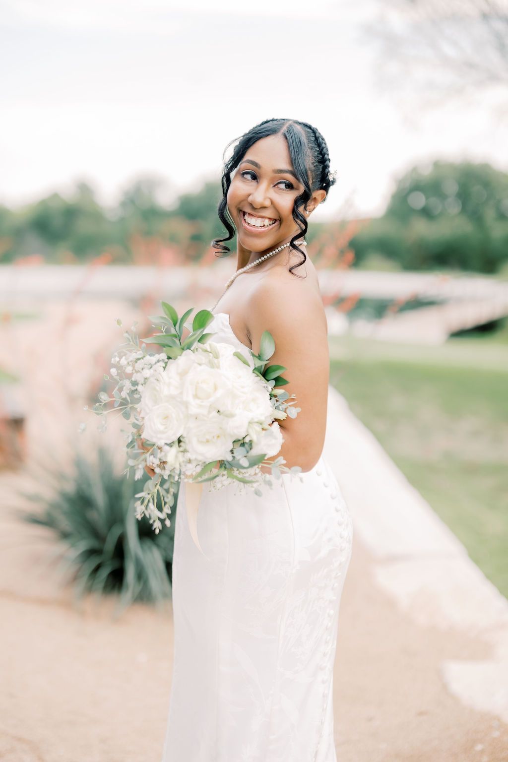 Smiling bride in white strapless gown holds white floral bouquet with greenery, posing outdoors on her wedding day.