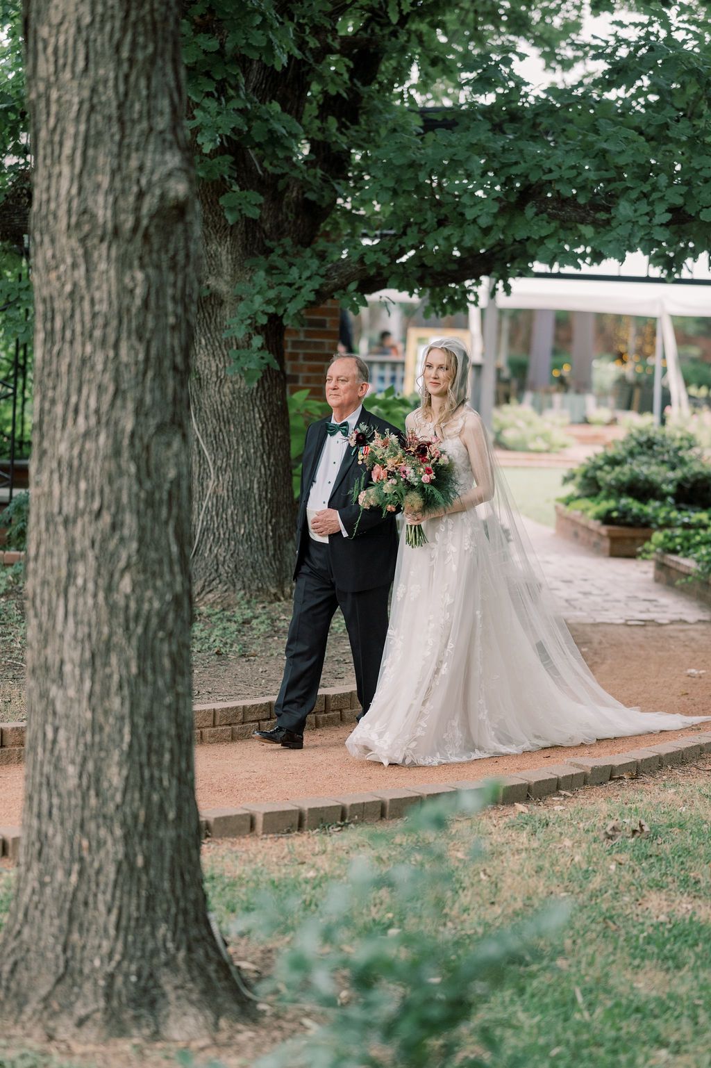 Bride in white gown holds bouquet while walking with escort through garden path under large oak tree.
