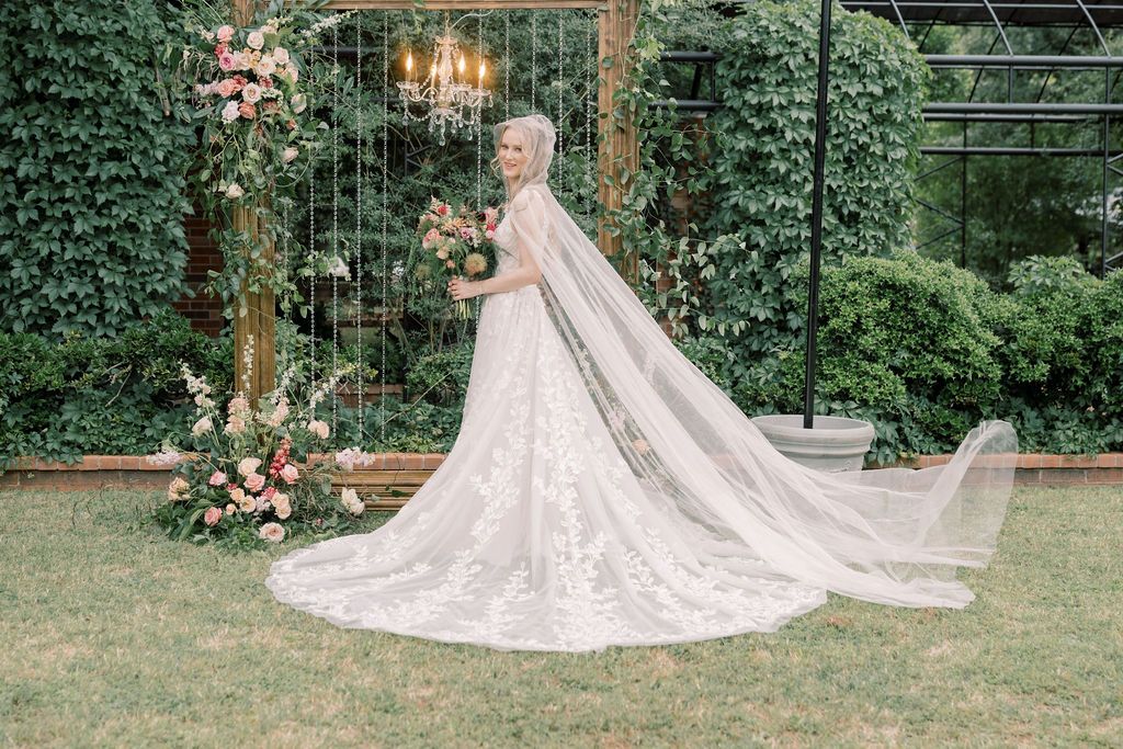 Bride in lace wedding gown with long cathedral veil stands in garden before floral arch with pink roses.