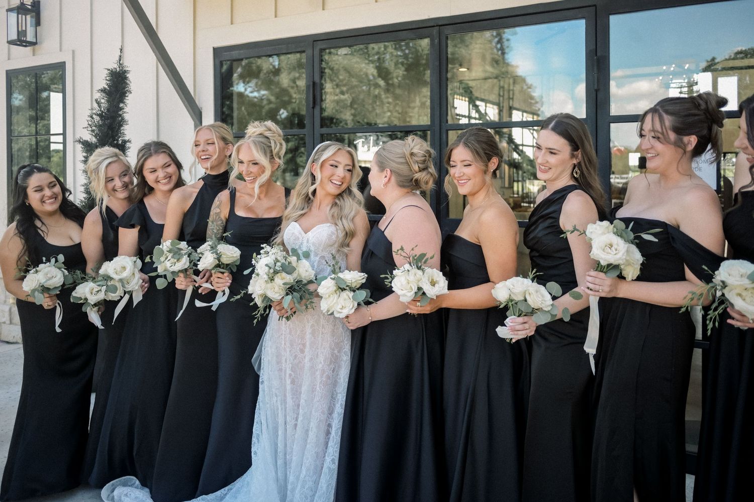 Bride poses with bridesmaids in black gowns holding white bouquets outside a modern wedding venue.
