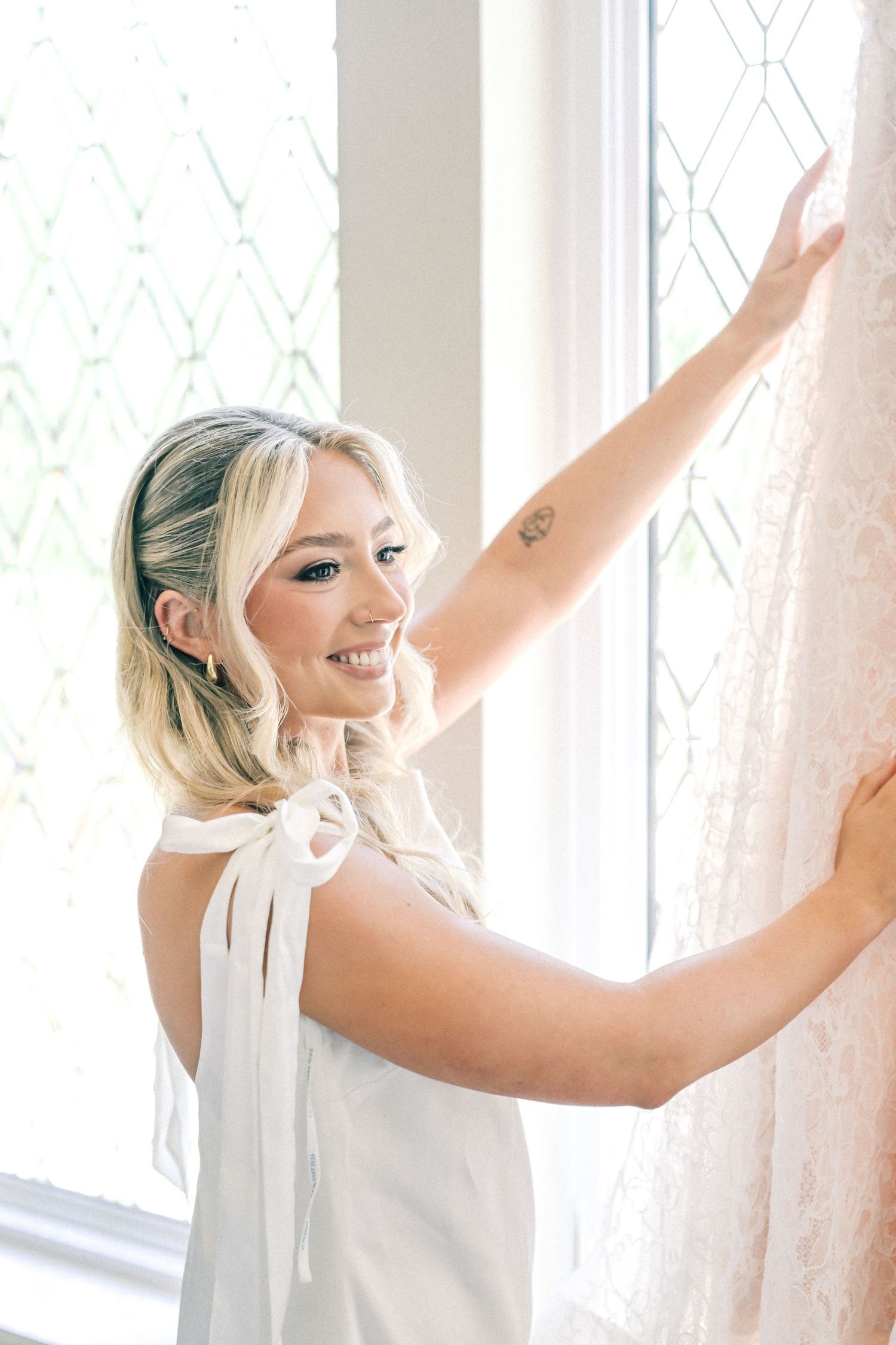 Smiling blonde bride in white robe touches lace wedding dress hanging by decorative window.