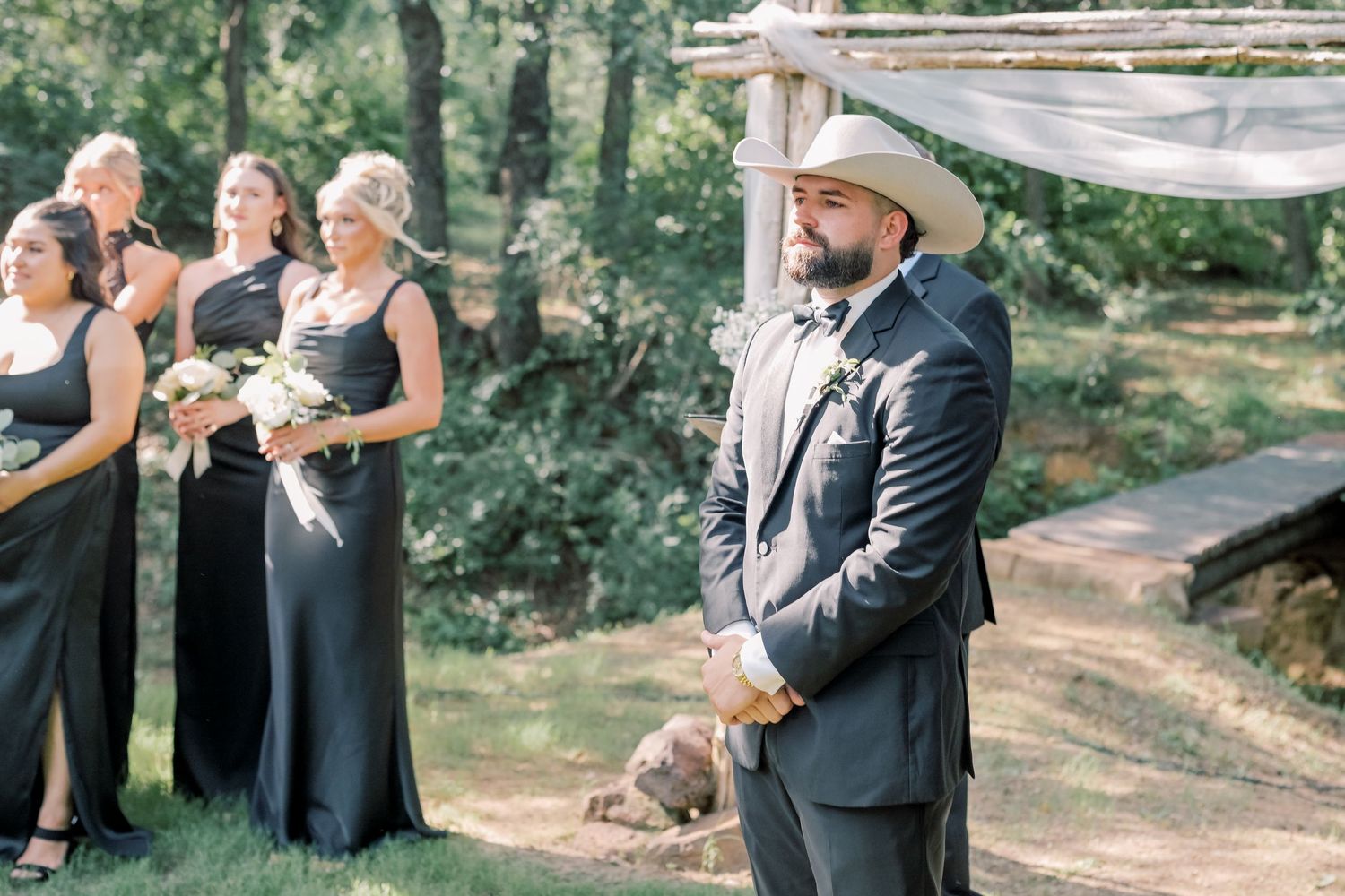 Groom in black suit and white cowboy hat waits at rustic altar flanked by bridesmaids in black gowns.