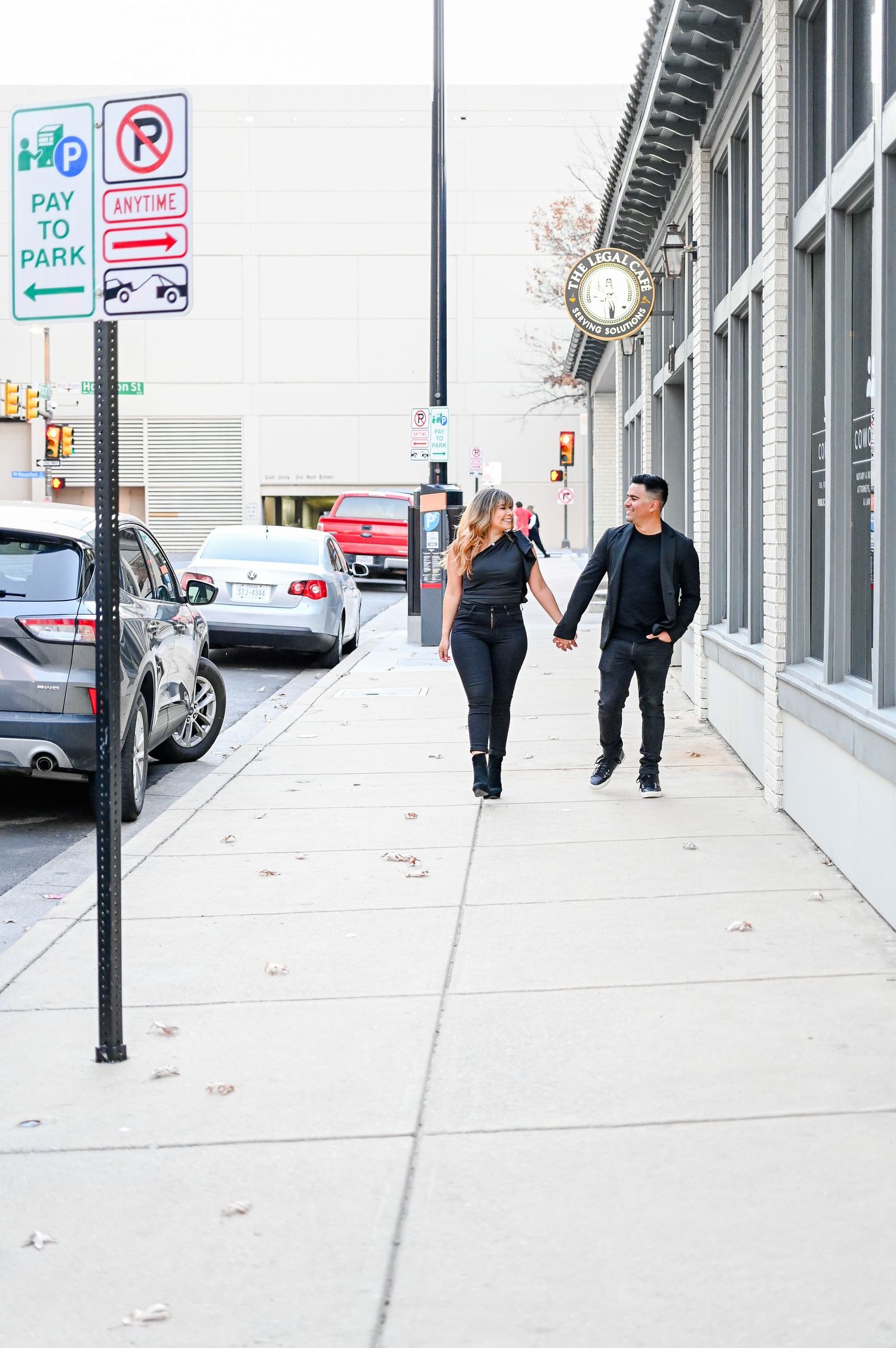 A couple dressed in black holds hands while walking together on an urban city sidewalk.