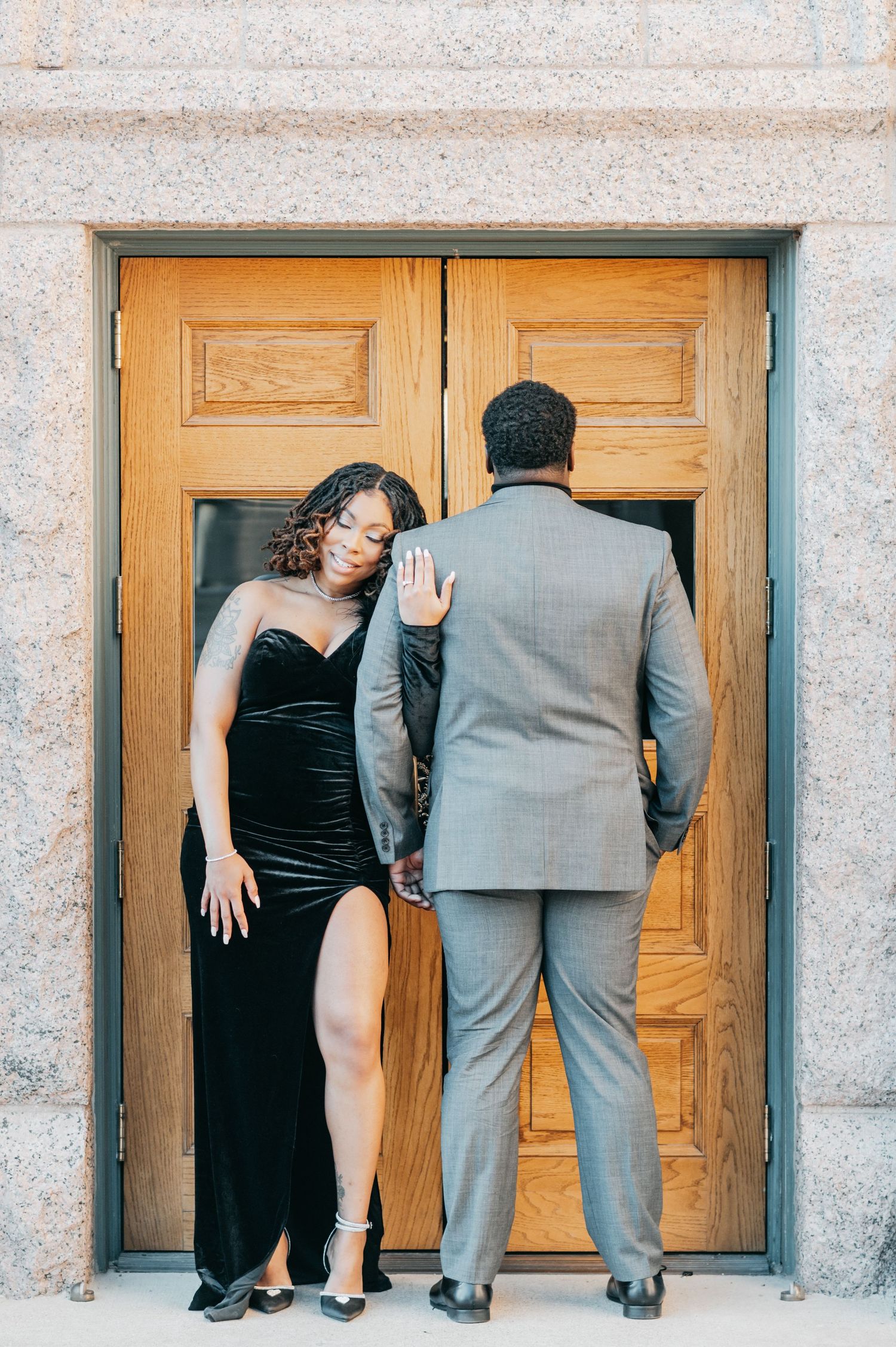 Couple posing by wooden doors, woman in black velvet gown and man in gray suit, elegant engagement photo.