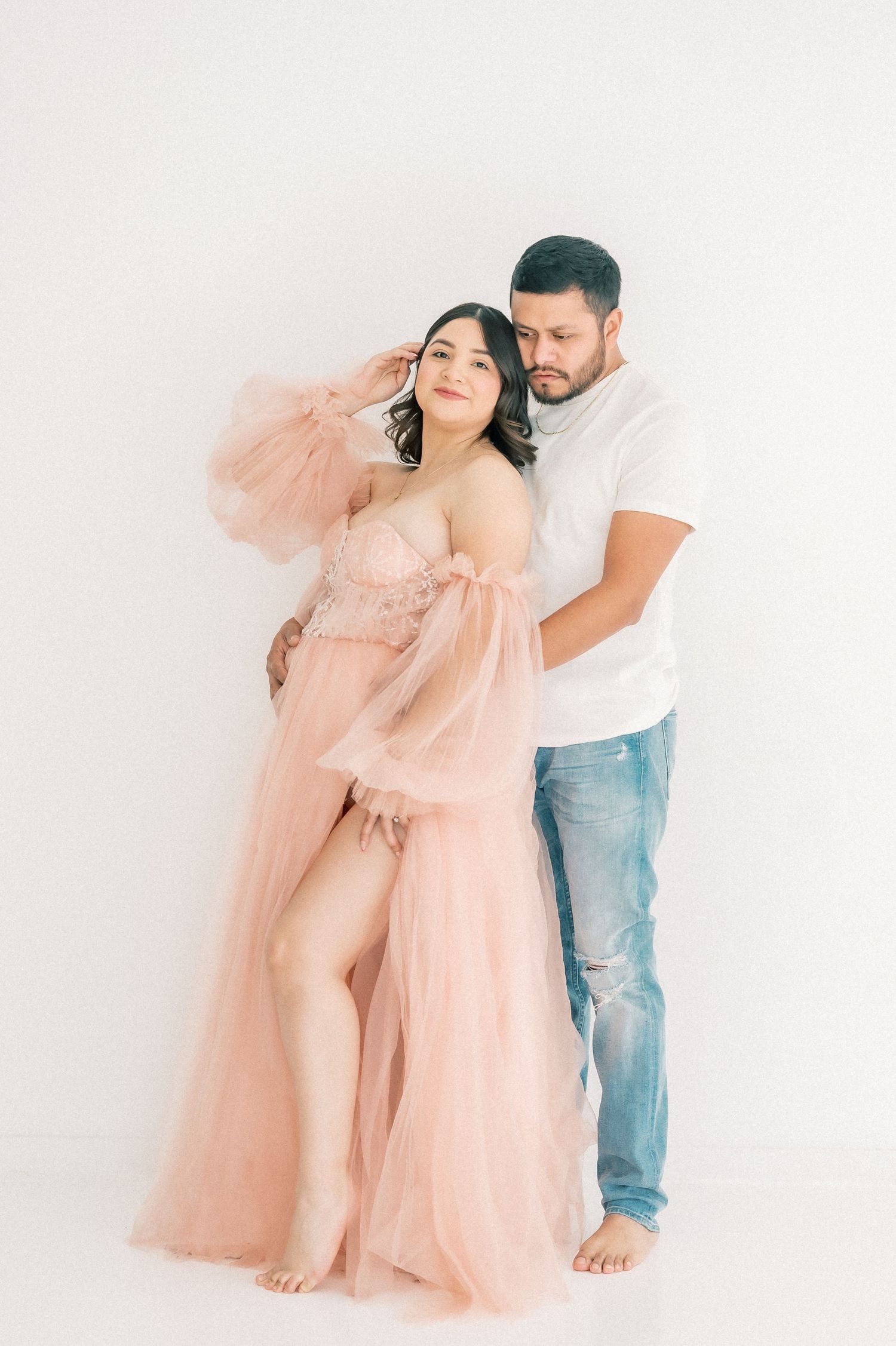 Couple maternity photo, woman in pink tulle gown embraced by man in white tee.