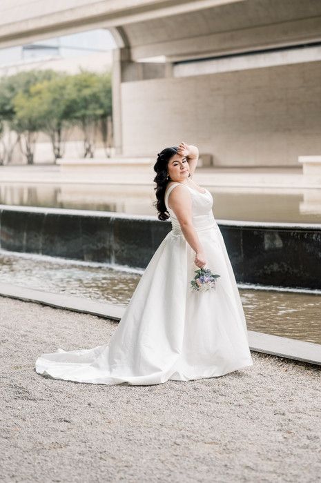 Bride in a white ball gown holding a bouquet poses gracefully near a reflective fountain outdoors.