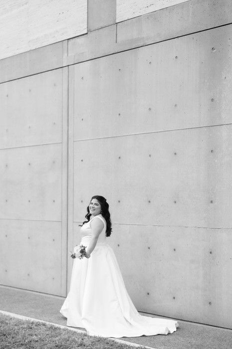 Black and white photo of a smiling bride in a white gown holding a bouquet against a concrete wall.
