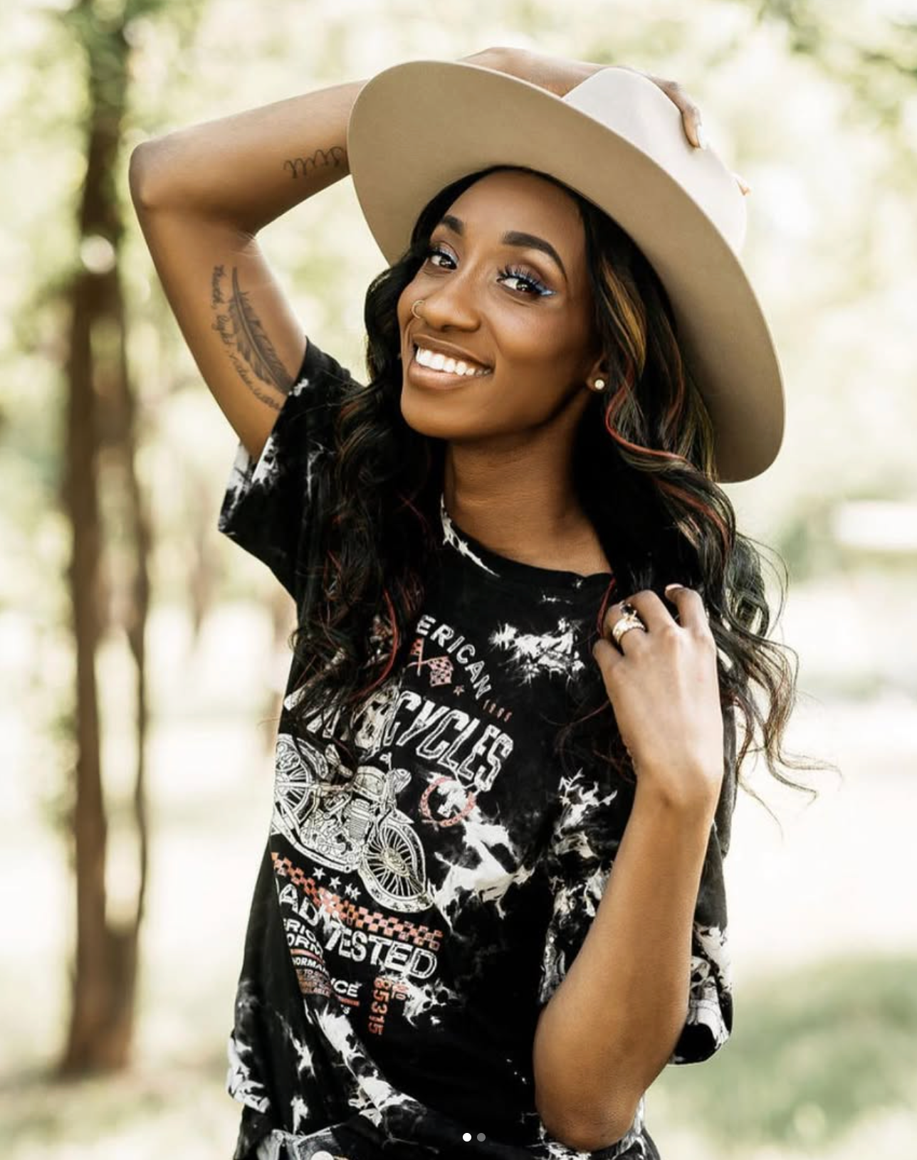 Smiling woman wearing a wide-brim tan hat and black graphic tee, posing outdoors with lush green trees in the background.