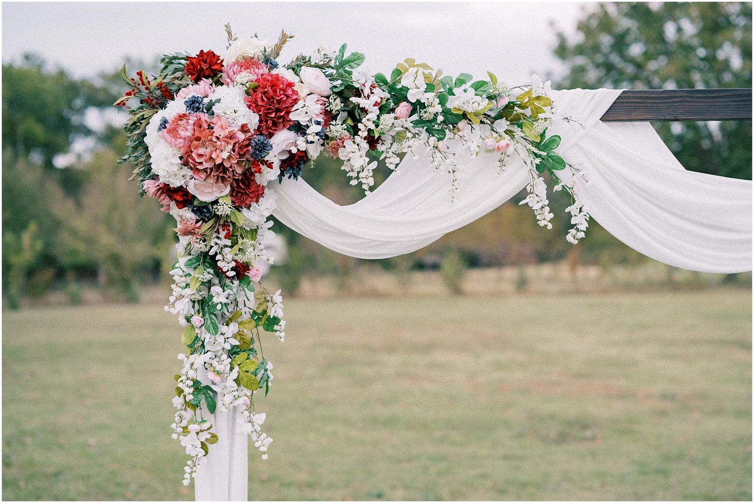 Floral wedding arch with white fabric and pink blooms against an outdoor backdrop.