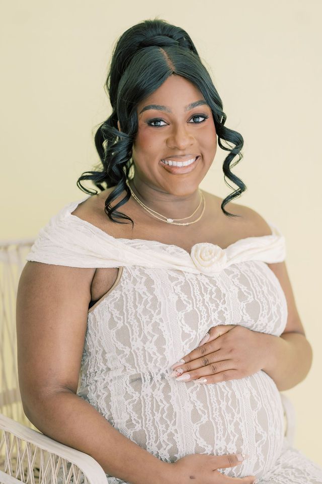 Smiling pregnant woman in white lace off-shoulder gown cradling baby bump during elegant maternity photo session.