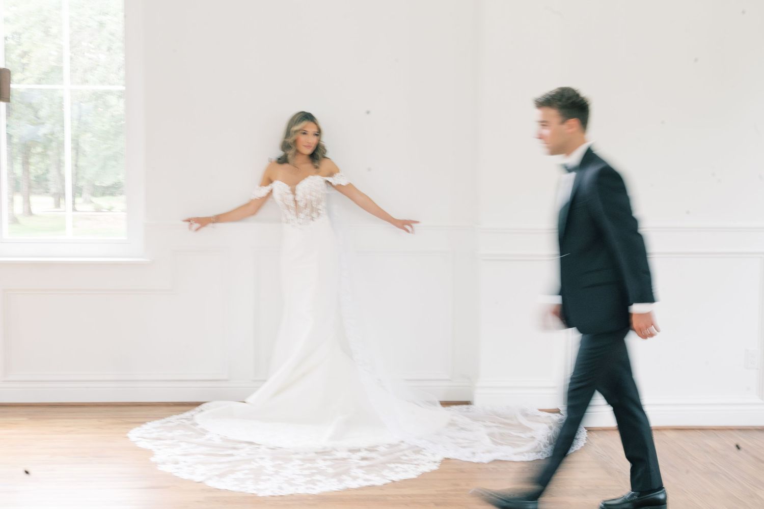 Bride in lace mermaid gown spreading arms as groom walks toward her in bright white room.