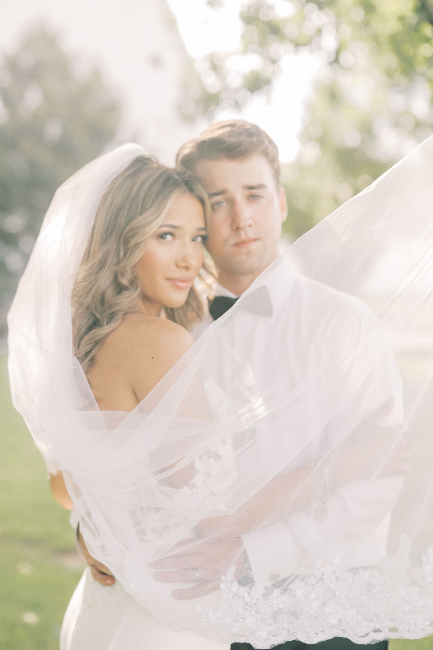 Bride and groom wrapped in flowing veil outdoors, soft golden light creating dreamy romantic portrait.