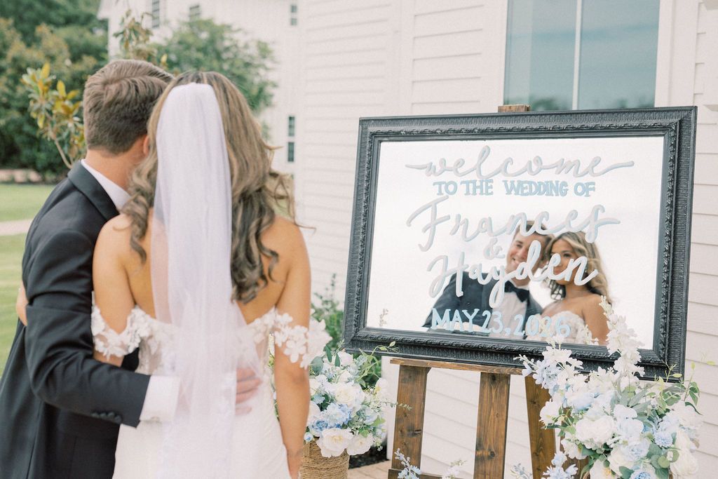 Bride and groom viewing elegant framed wedding welcome mirror sign on easel with floral arrangements at outdoor ceremony.