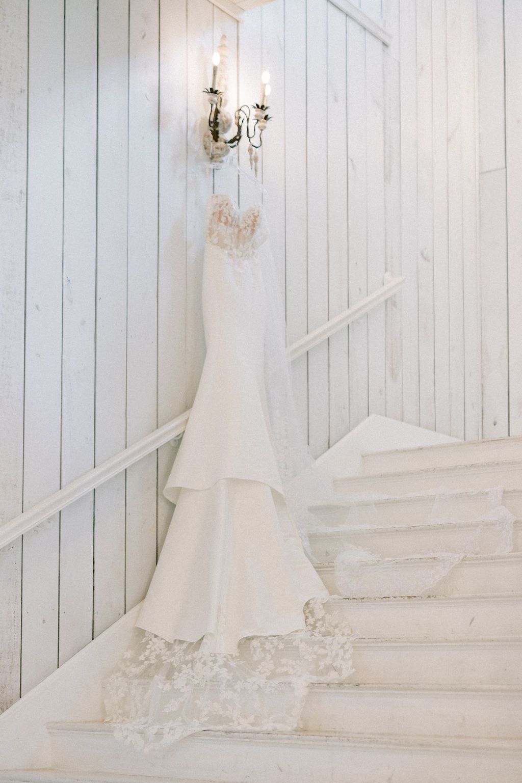 Elegant mermaid wedding dress with lace hem hanging on white wooden staircase beside candle sconce.