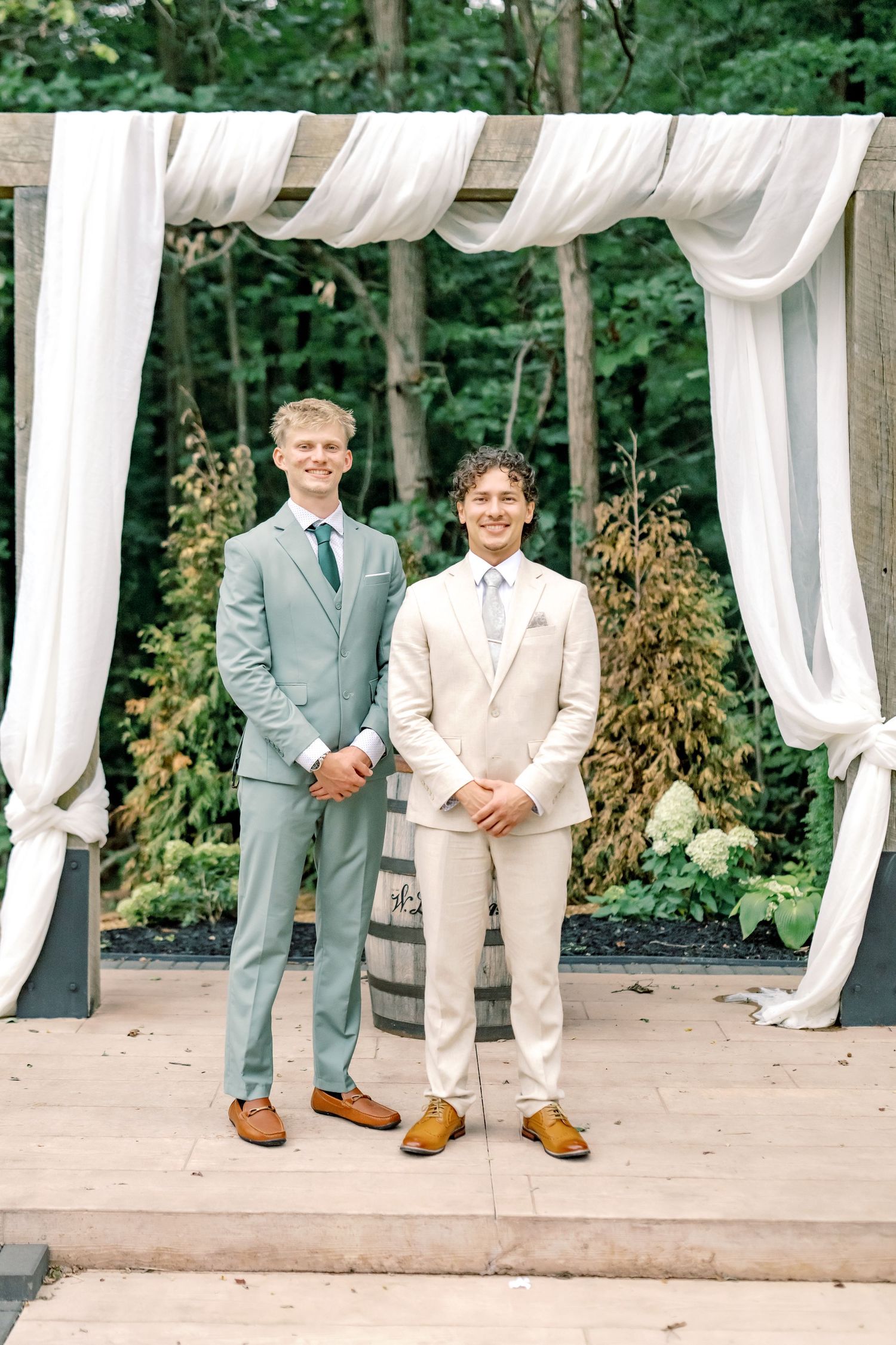 Two grooms in light suits stand smiling under a draped white arch at an outdoor wedding ceremony.