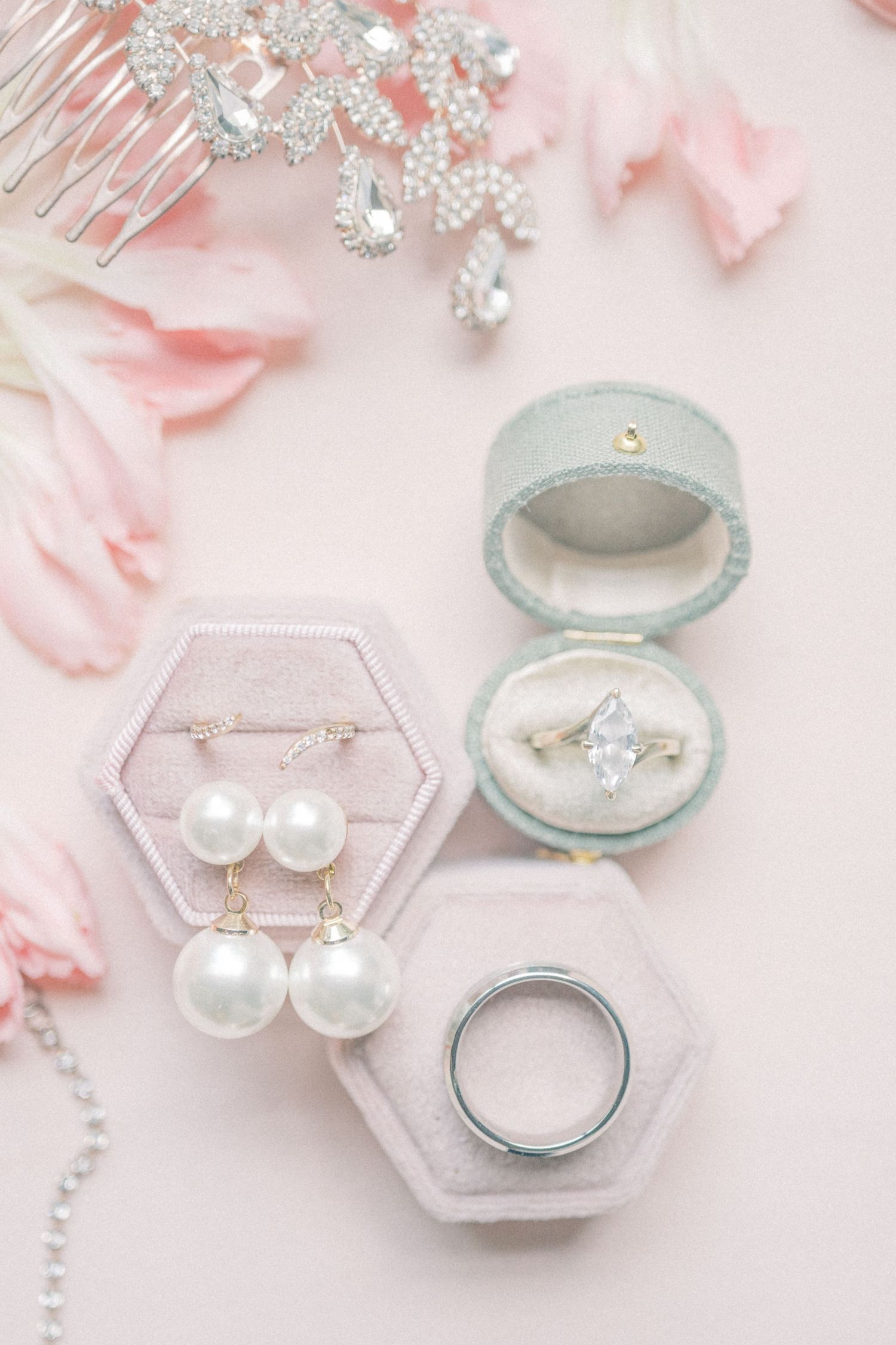 Elegant bridal jewelry flat lay with pearl drop earrings, diamond engagement ring, wedding band, and crystal hair comb on blush pink.
