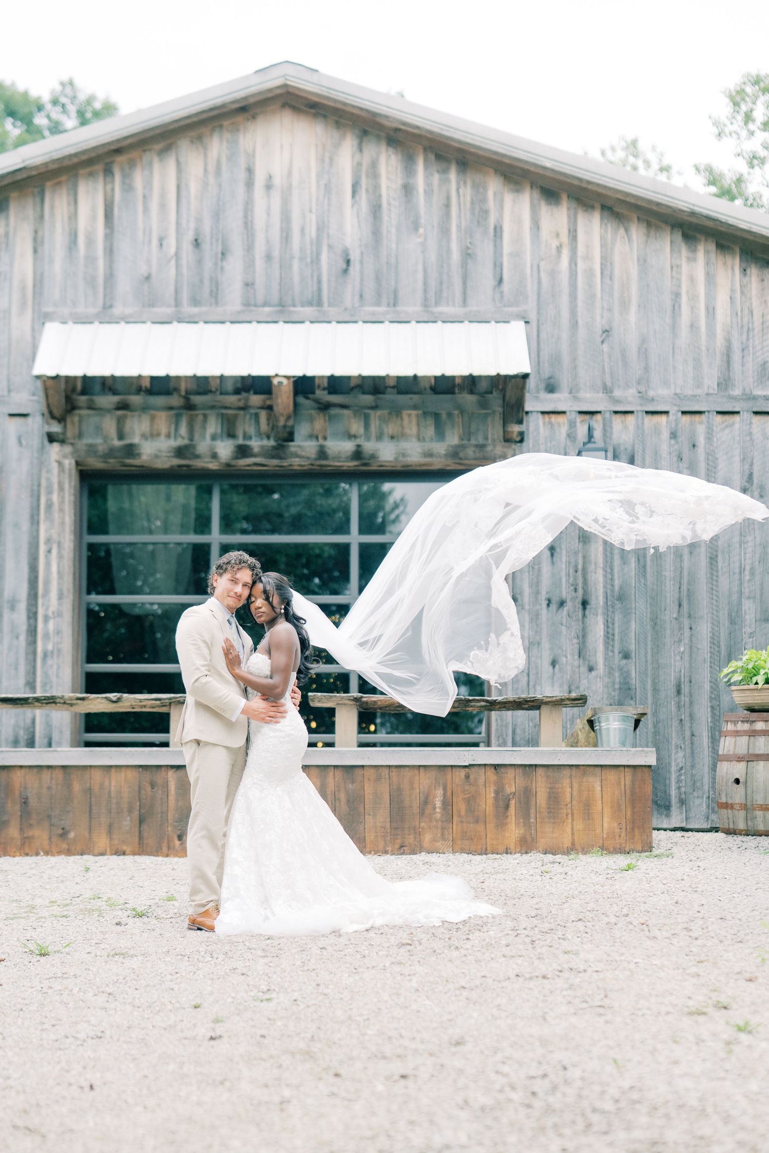Bride and groom pose together as wedding veil dramatically flows in wind before rustic barn backdrop.