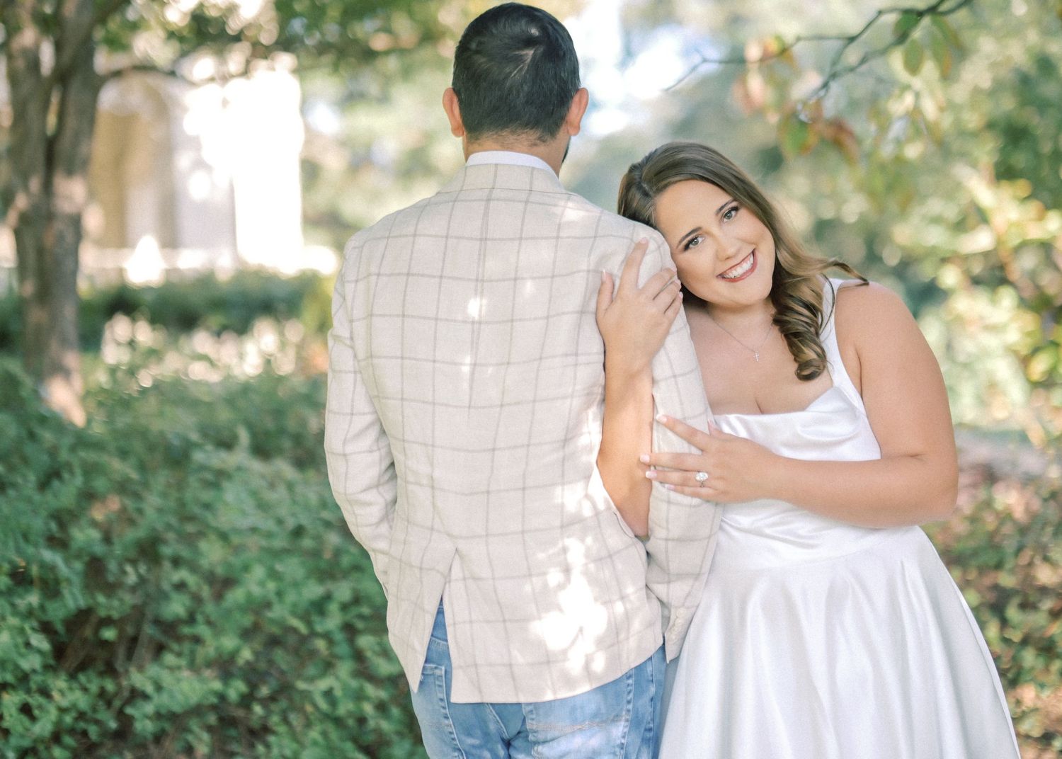 Smiling woman in a white dress leans against a man in a plaid blazer facing away in a sunlit garden.