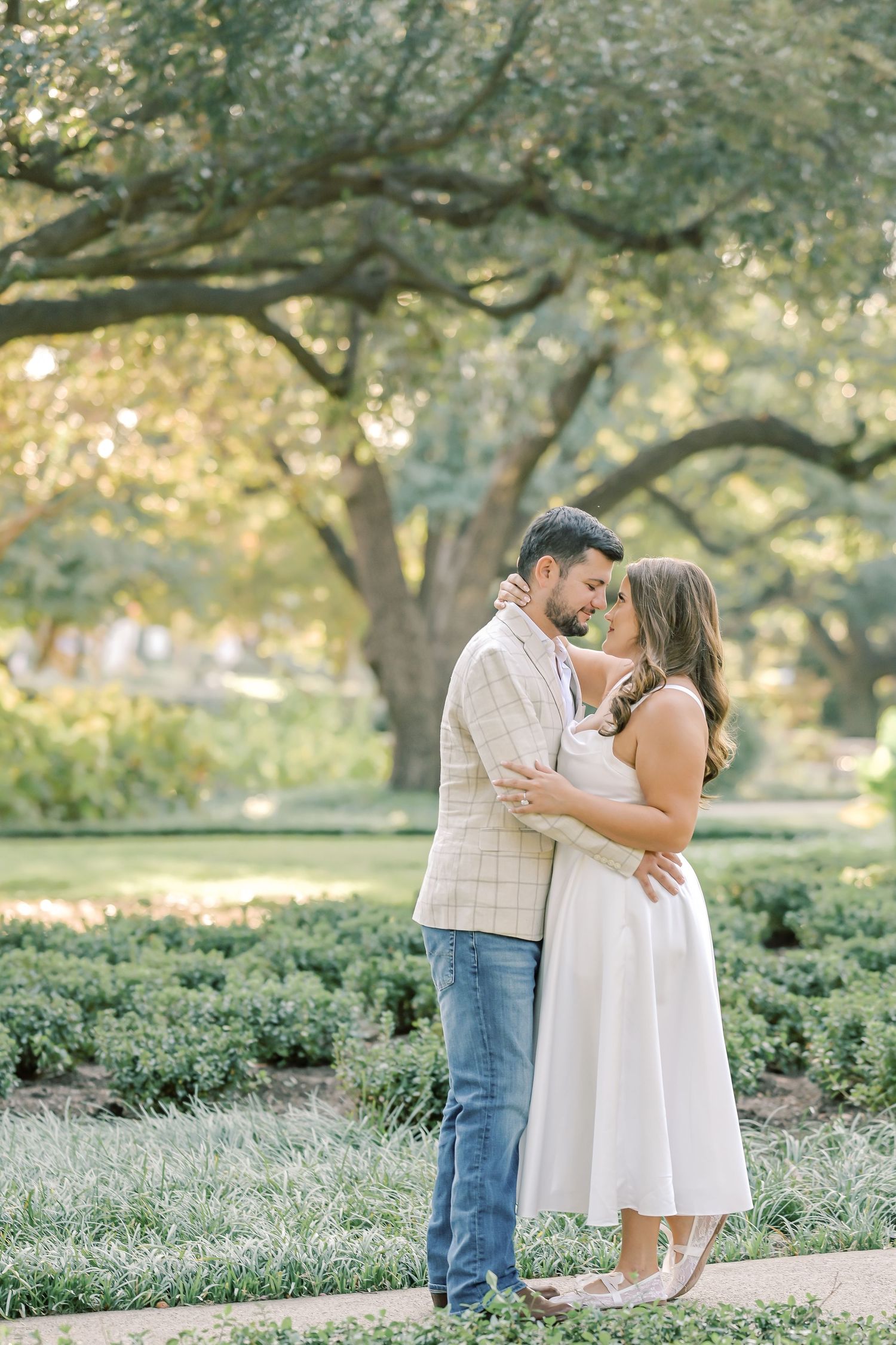 Couple in an affectionate embrace during an outdoor engagement photo session in a lush garden beneath large oak trees at golden hour.