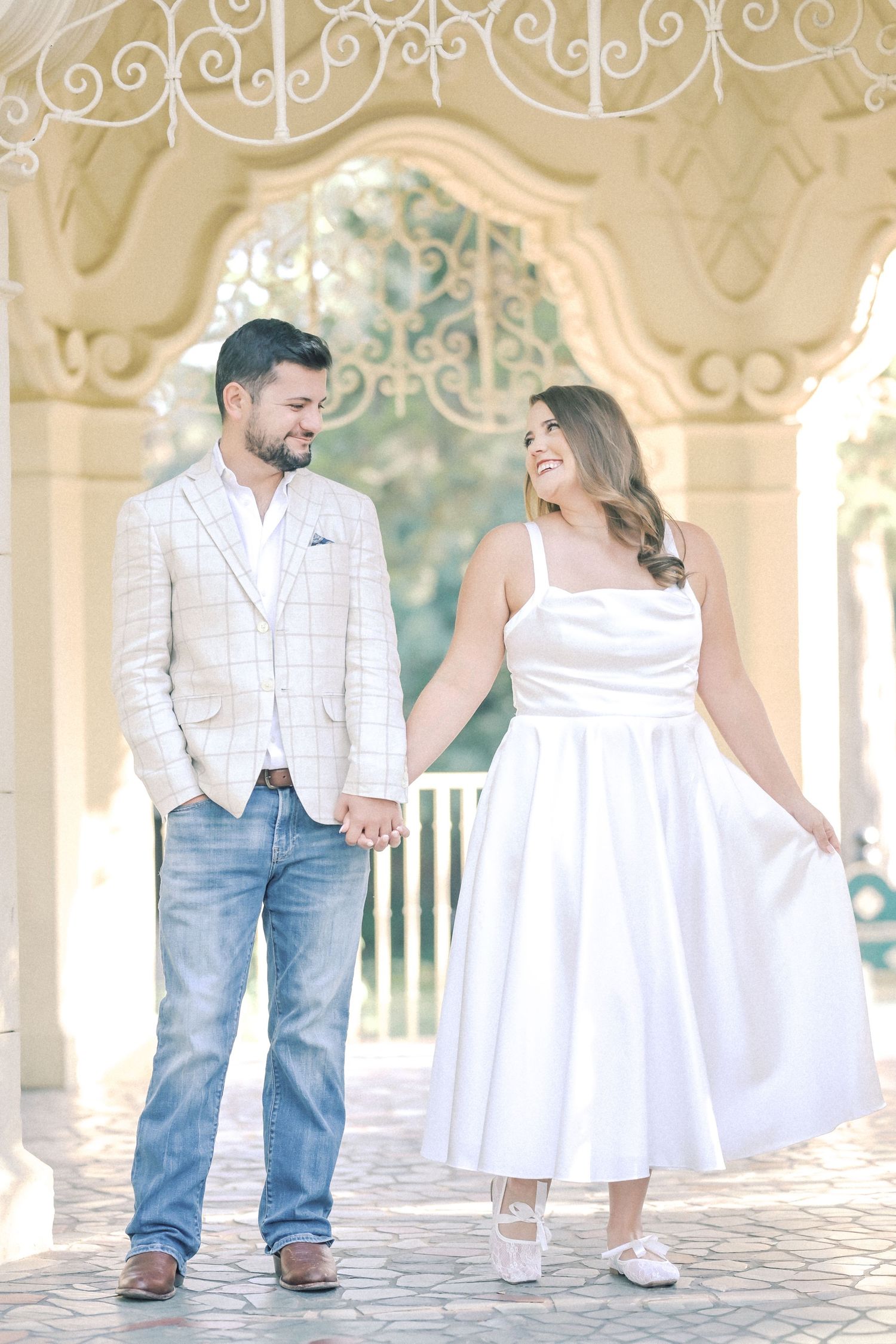 Smiling couple holding hands under an ornate white pavilion arch, he in a plaid blazer and jeans, she in a white satin dress.