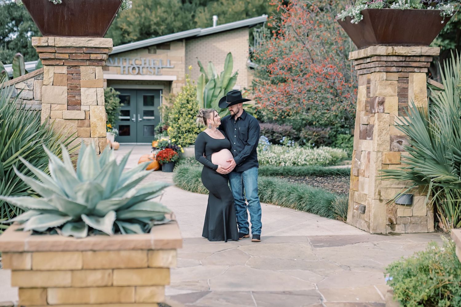 Pregnant woman in a black dress and man in a cowboy hat pose on a stone garden path with agave plants and fall foliage.