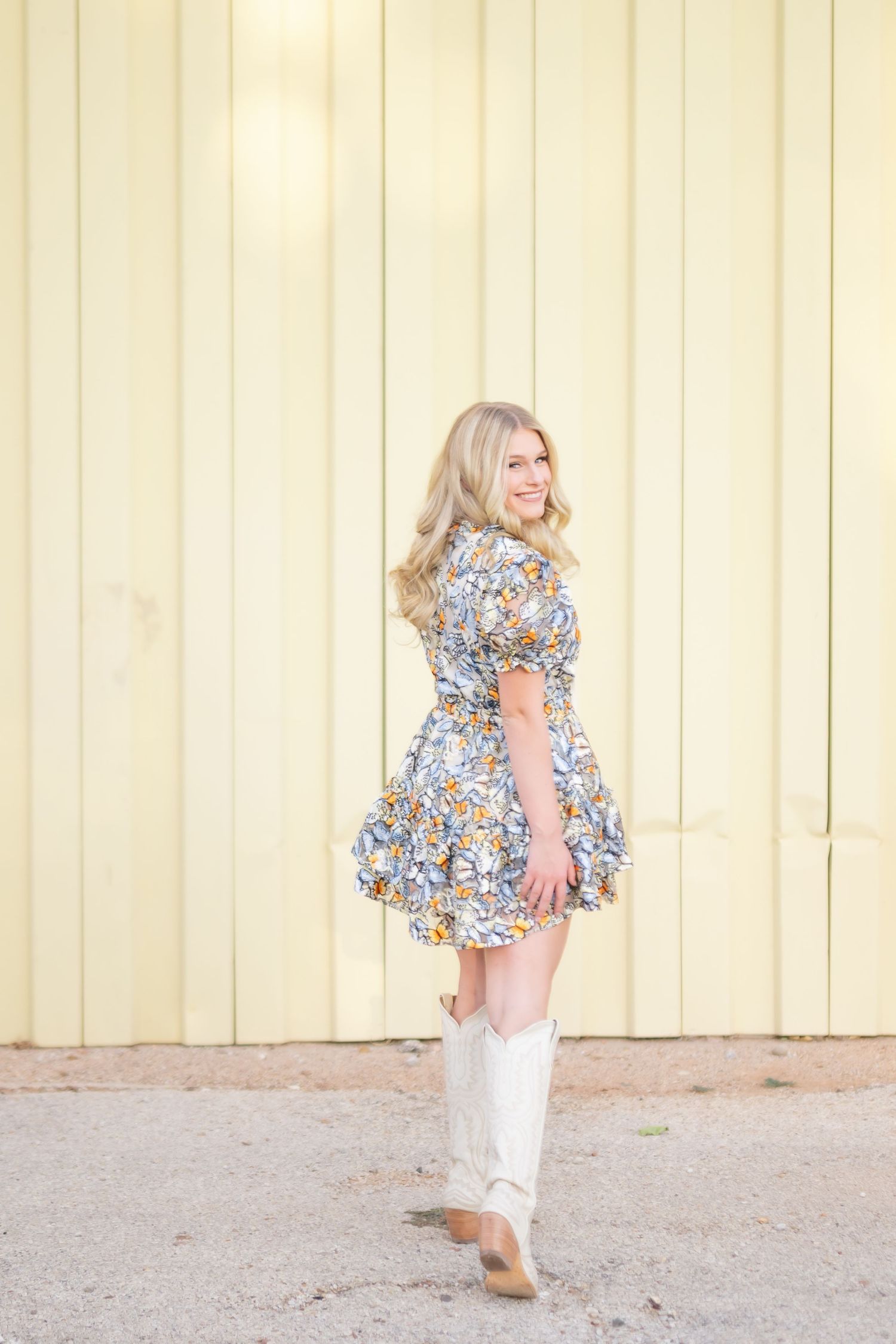 Blonde woman in floral mini dress and white cowboy boots smiling outdoors near yellow wall.