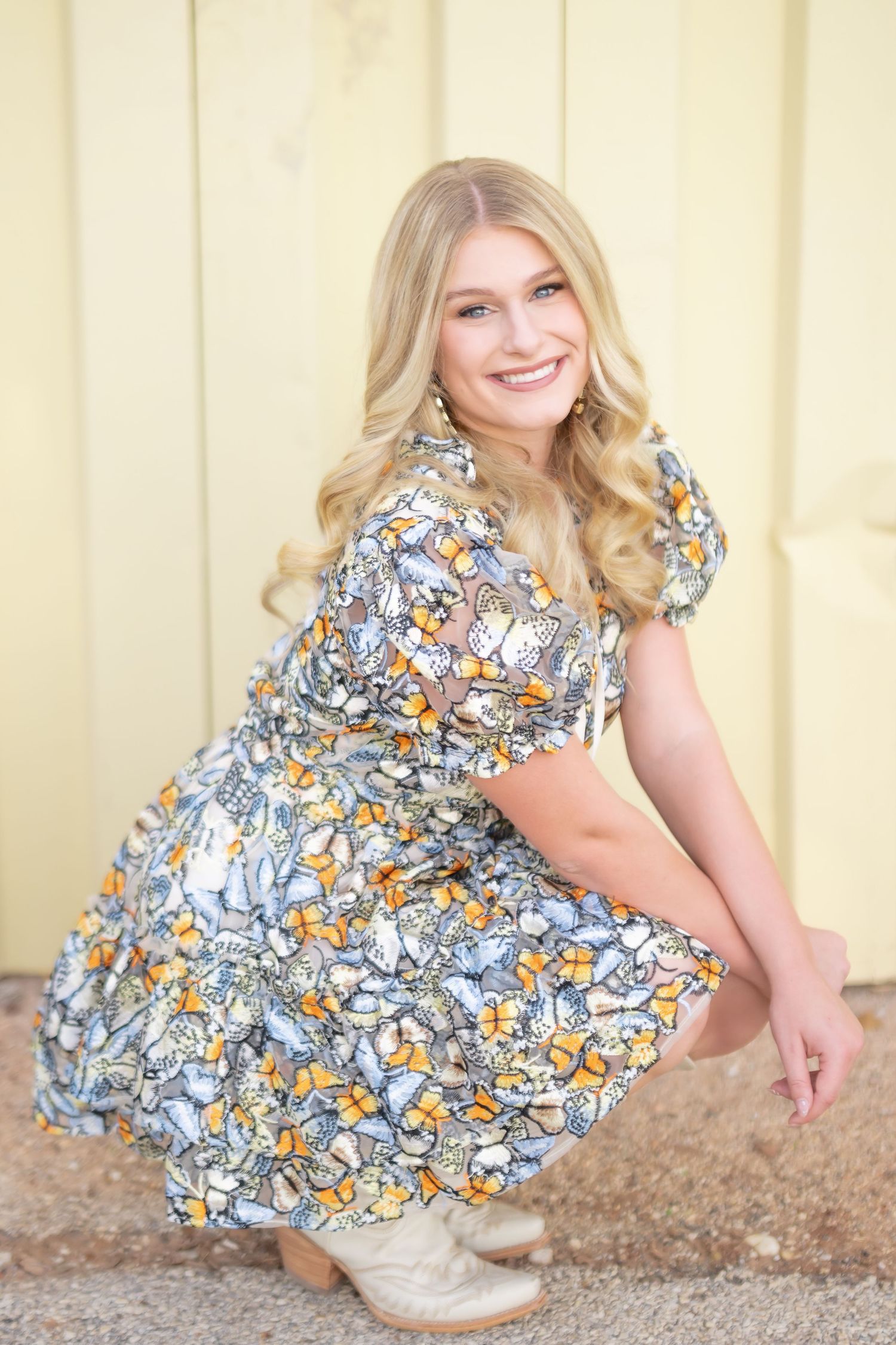 Smiling blonde woman in a floral butterfly dress and cowboy boots crouching outdoors near a yellow wall.