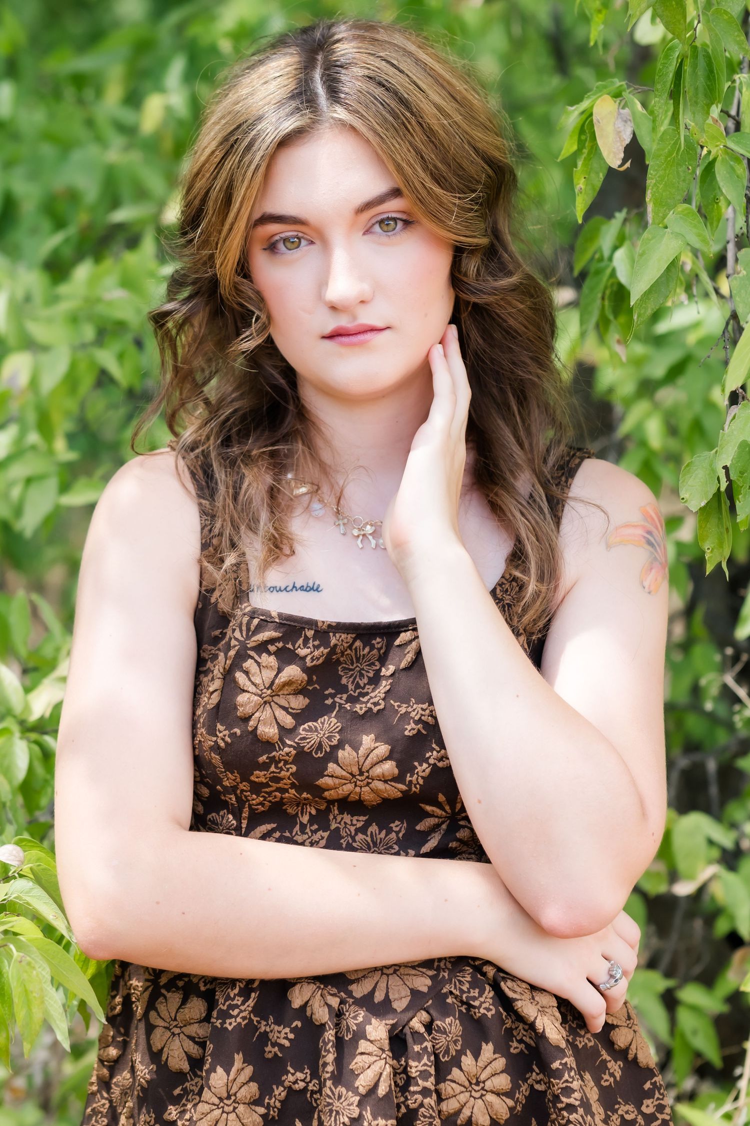 Young woman with wavy brown hair in floral dress poses outdoors among green leaves, hand touching her face.