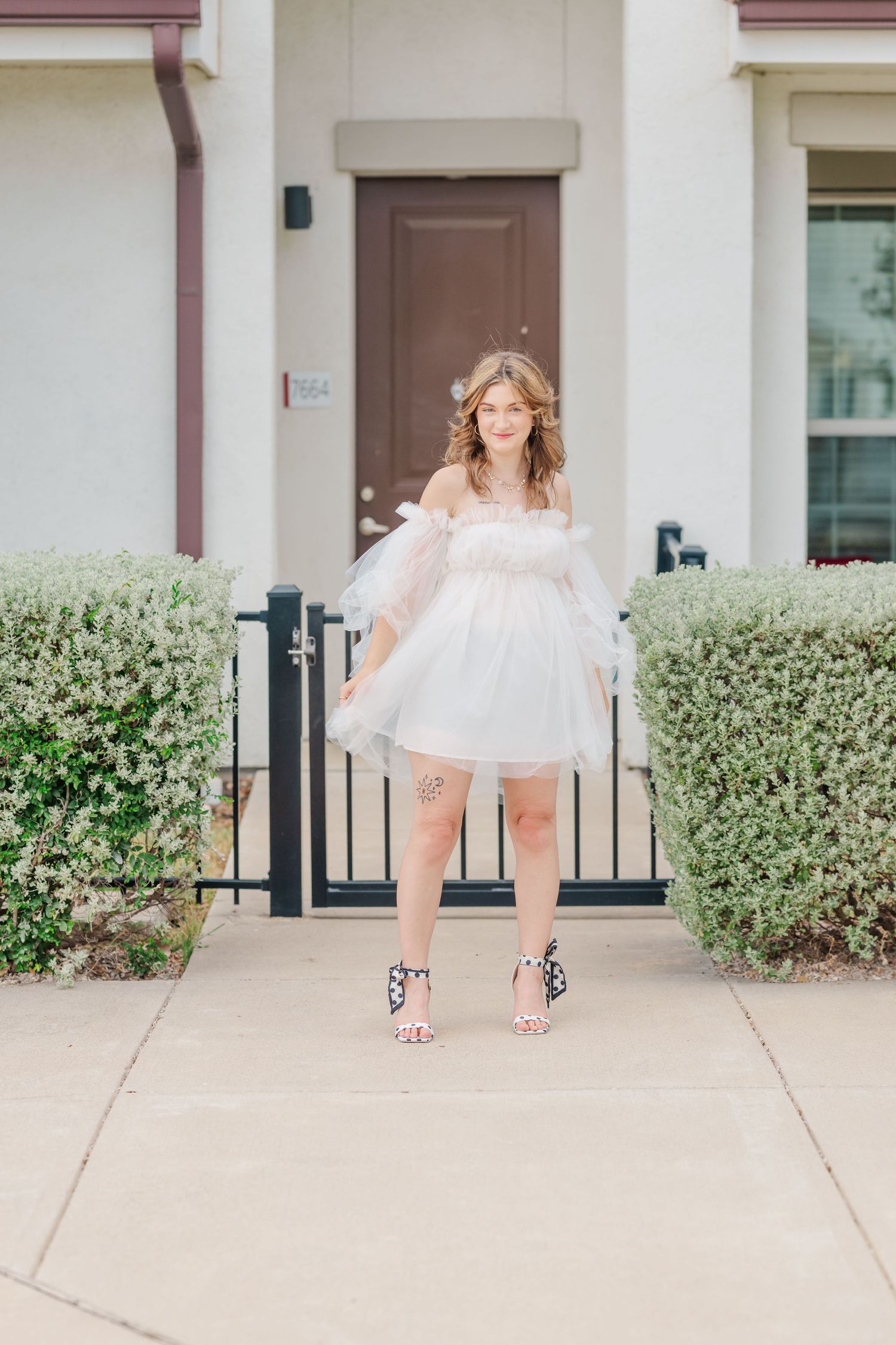 Woman in white tulle off-shoulder mini dress and heels posing on sidewalk in front of home entrance.