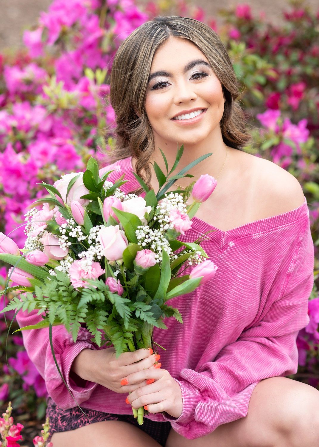 Young woman in pink off-shoulder sweater holding pink tulip bouquet, smiling outdoors among blooming pink azalea bushes.