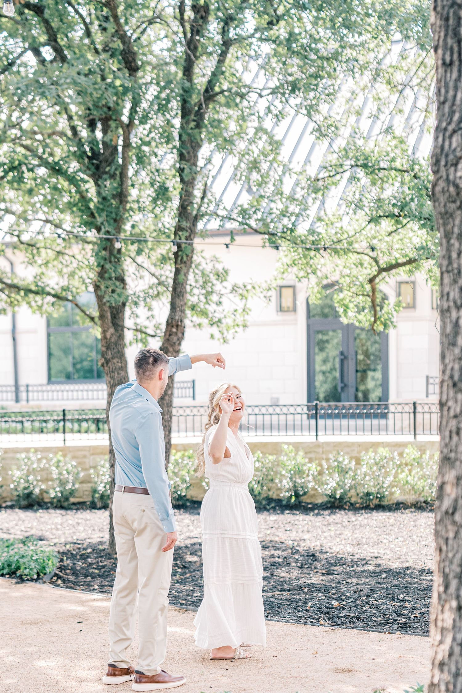 A couple dances outdoors near trees, the man spinning the woman in a white dress in a sunlit garden.