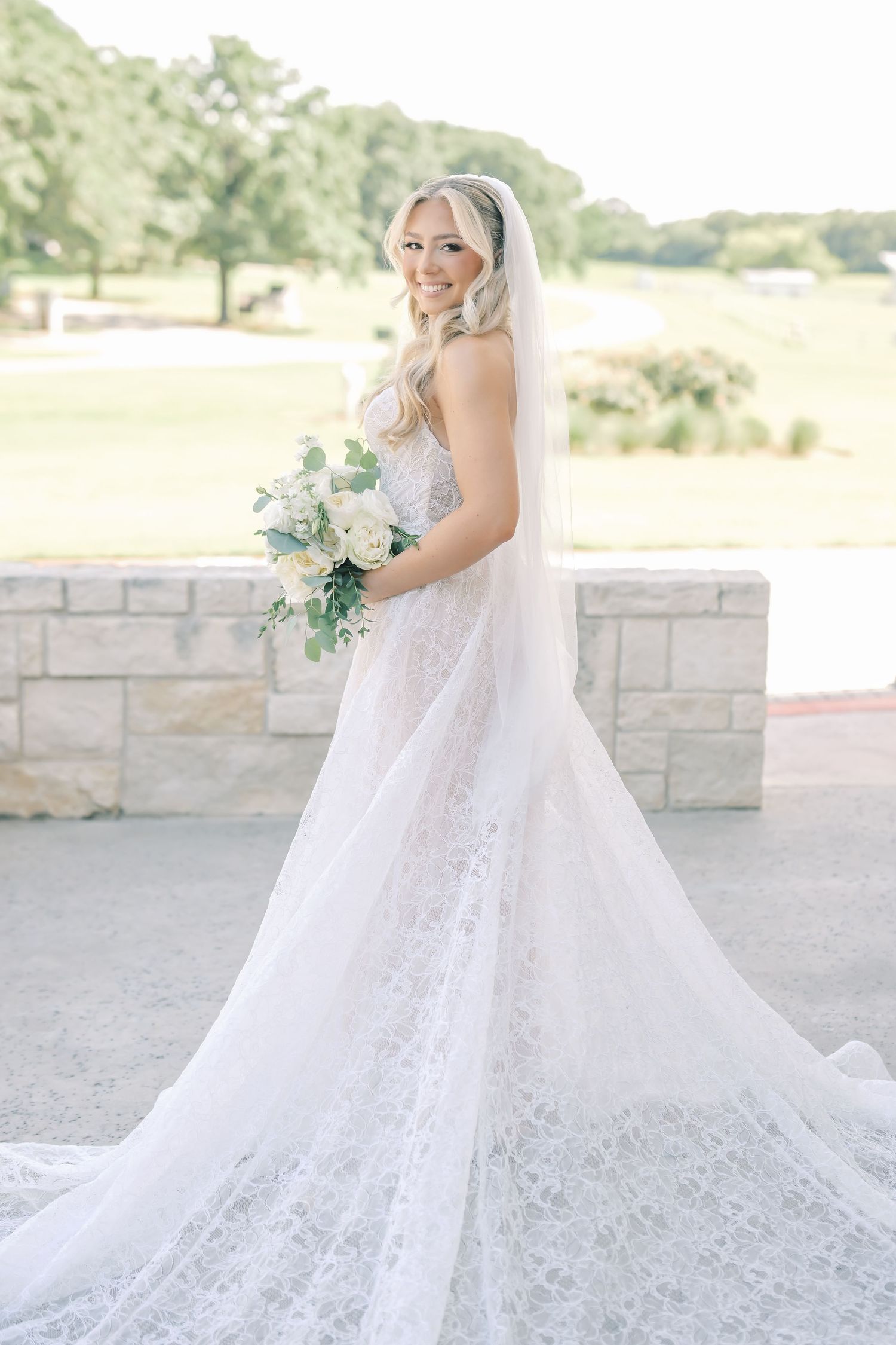 Elegant bride in lace wedding gown with cathedral veil holding white rose bouquet outdoors.