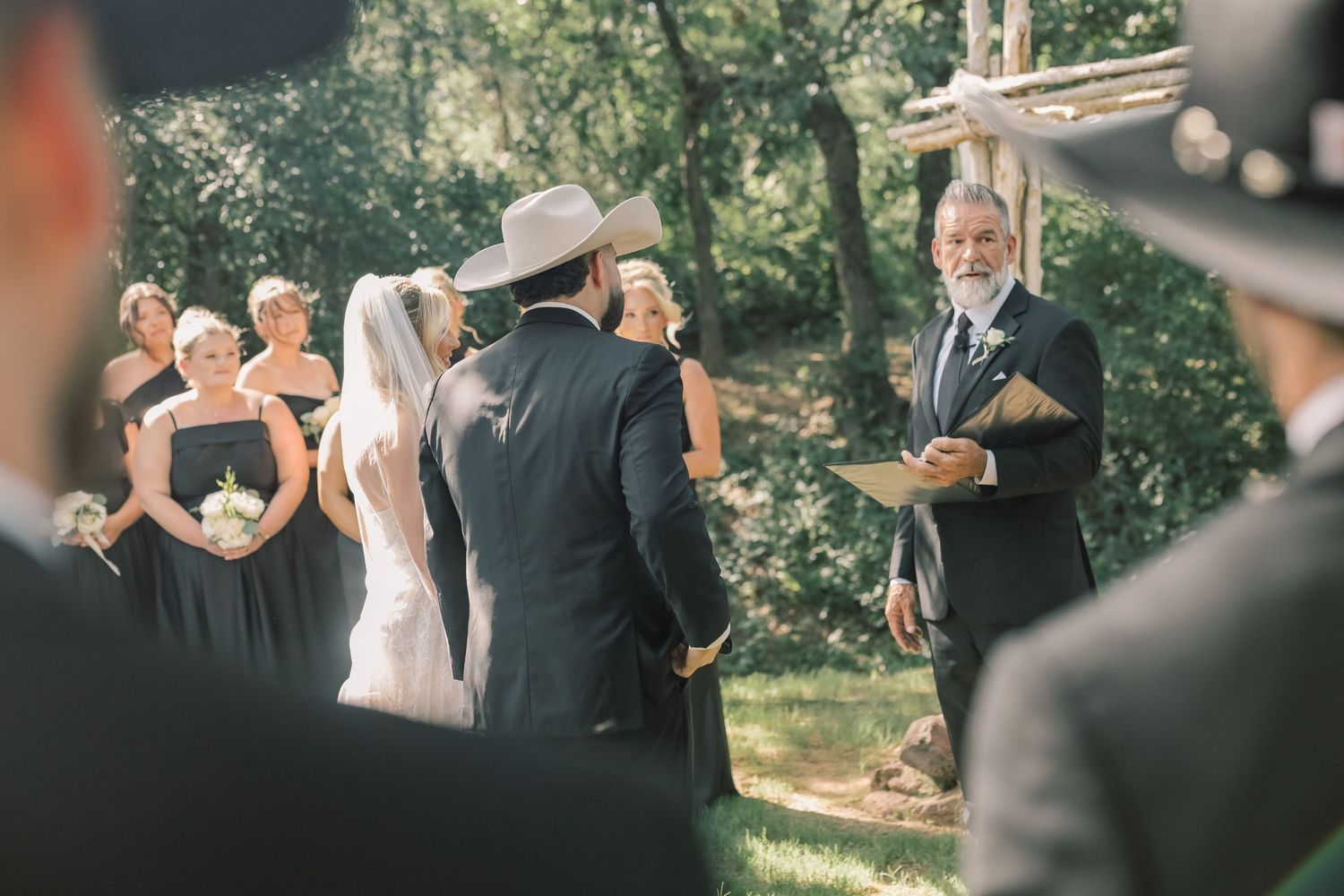 Outdoor wedding ceremony with bride, groom in cowboy hat, officiant, and bridesmaids.