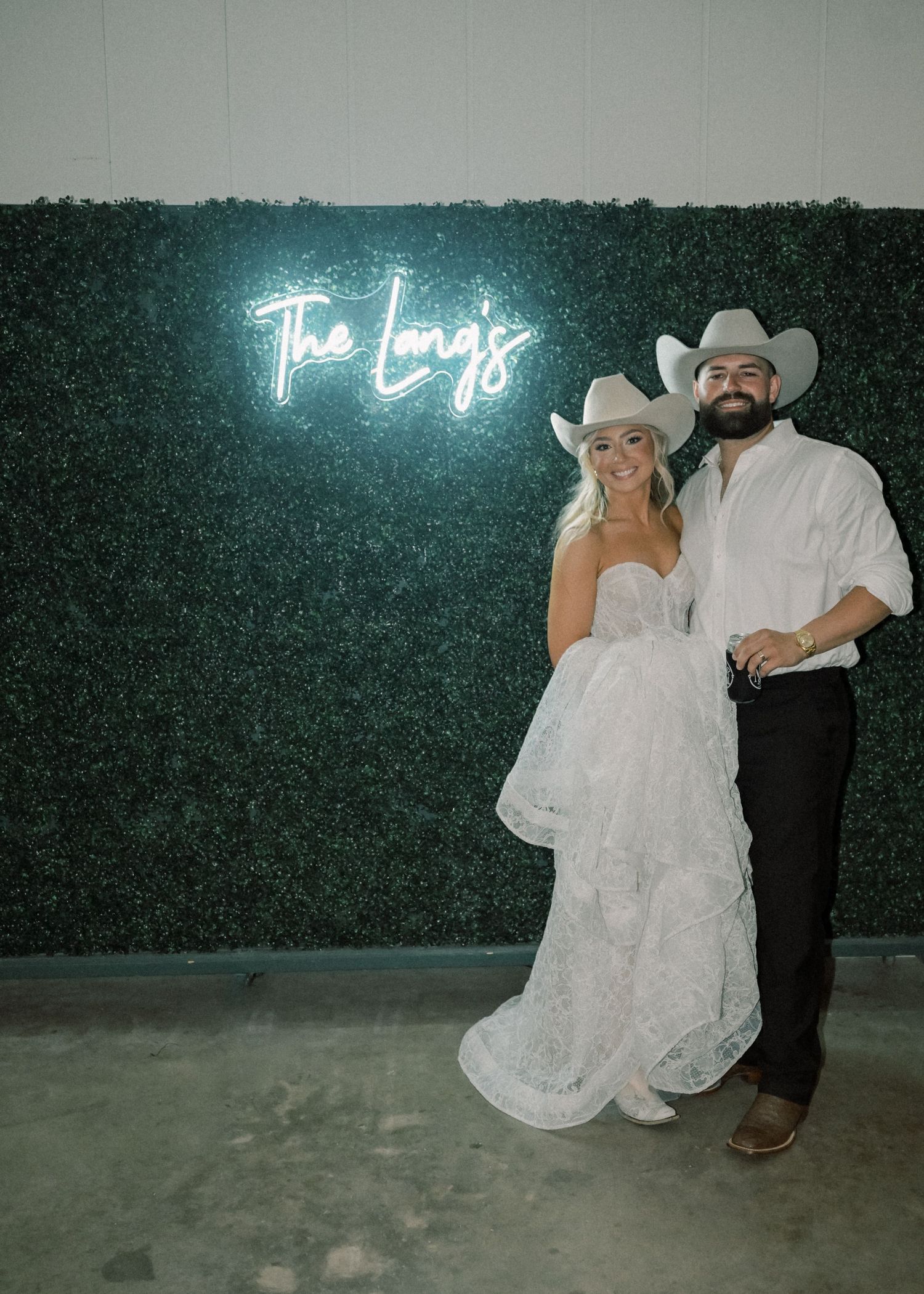 Newlywed couple in cowboy hats pose before green hedge wall with neon sign reading The Longs.