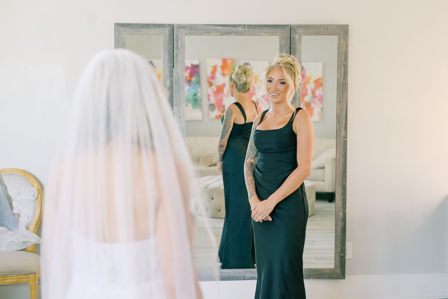 Bridesmaid in black gown reacting to bride in white veil during first look reveal in mirror.