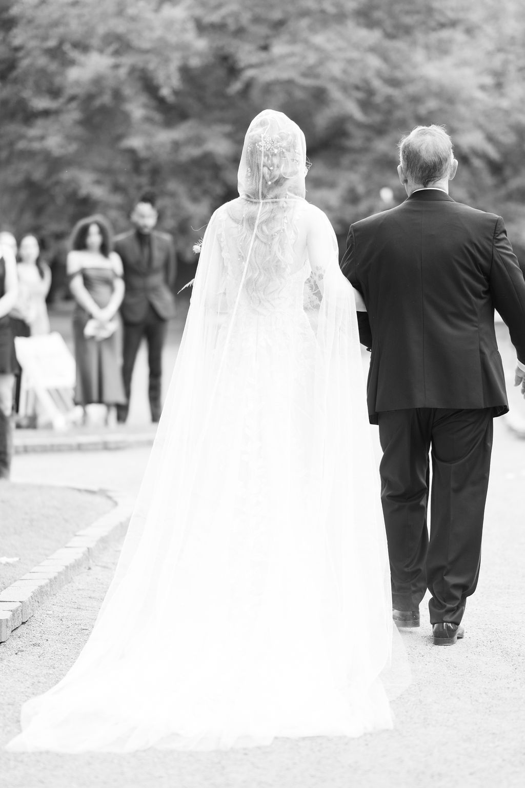Black and white photo of a bride in a lace veil walking down the aisle with her father at an outdoor wedding.