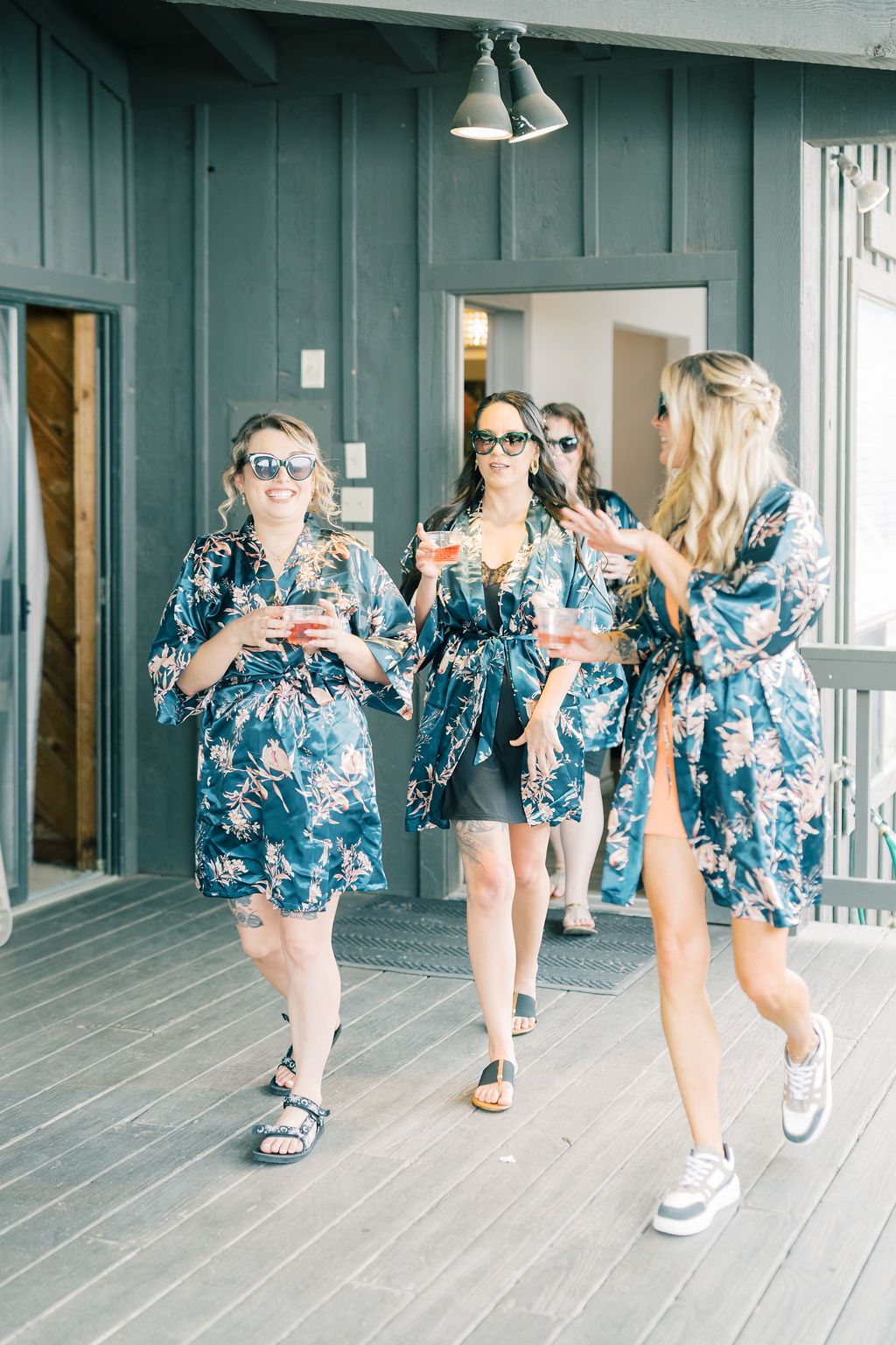 Bridesmaids in matching blue floral robes holding cocktails on a wooden deck during wedding preparations.