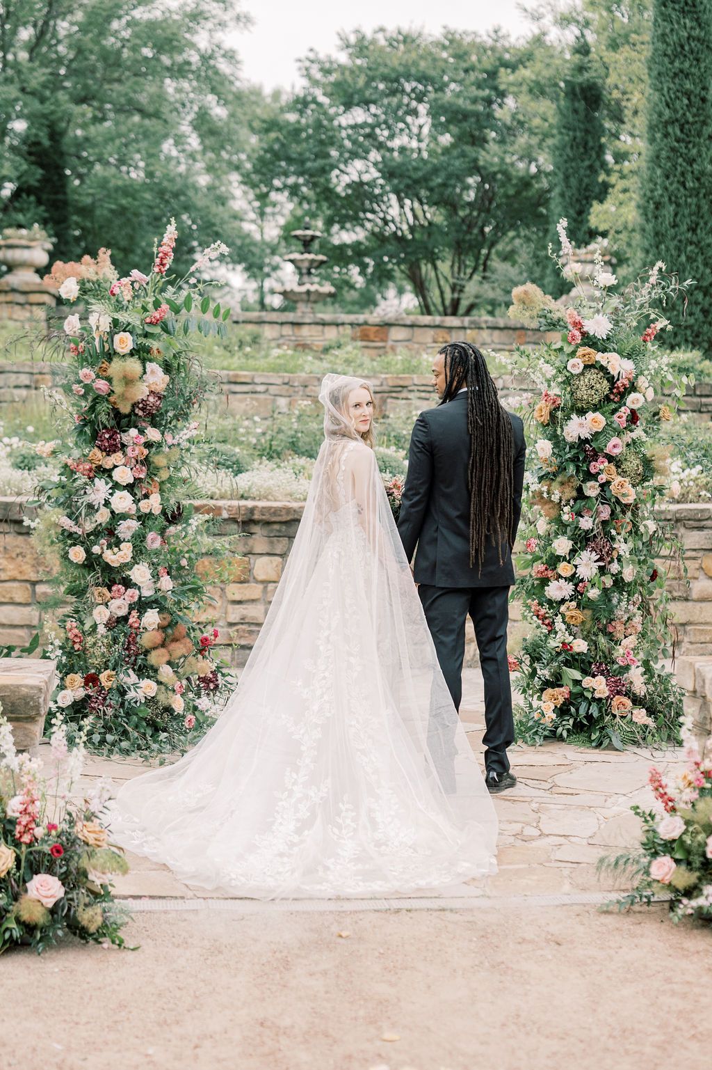 Bride in lace gown and veil stands with groom in black suit before floral arch in garden wedding ceremony.