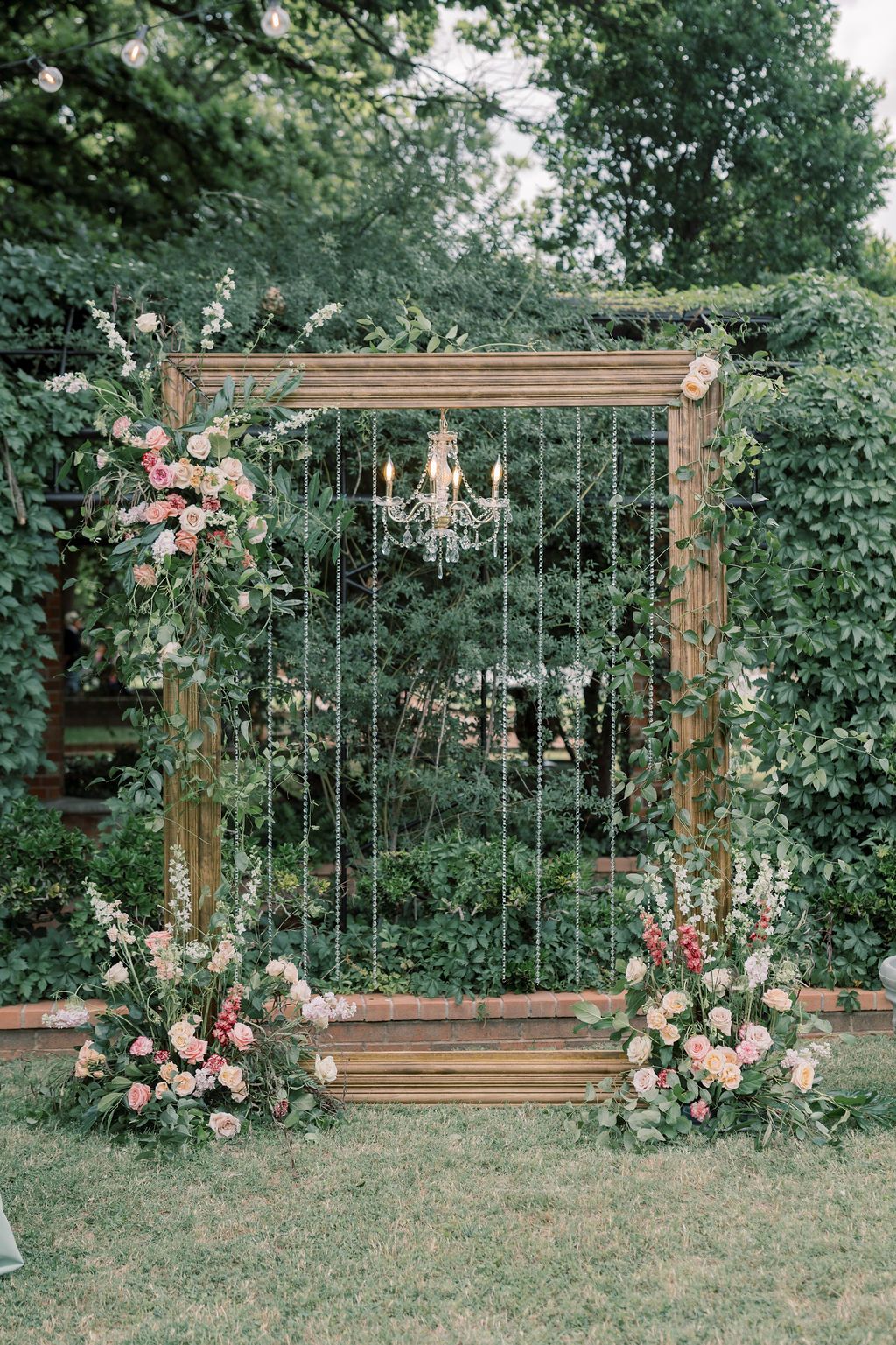 Rustic wooden frame wedding arch with chandelier, pink florals, and lush greenery in an outdoor garden setting.