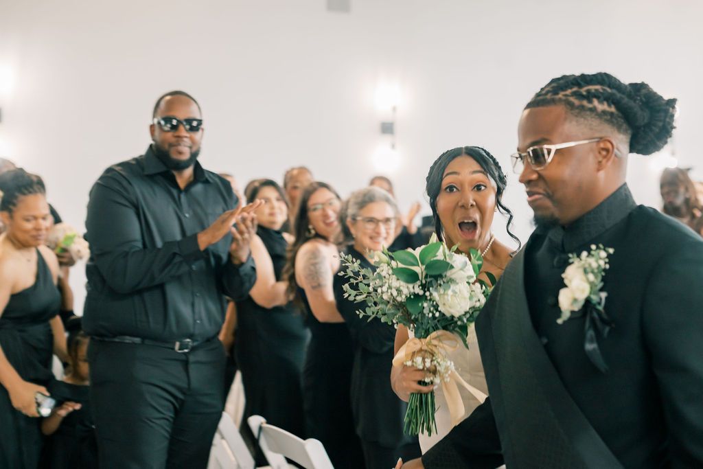 Joyful wedding ceremony moment with bride holding white bouquet, groom in black suit, and guests celebrating in background.