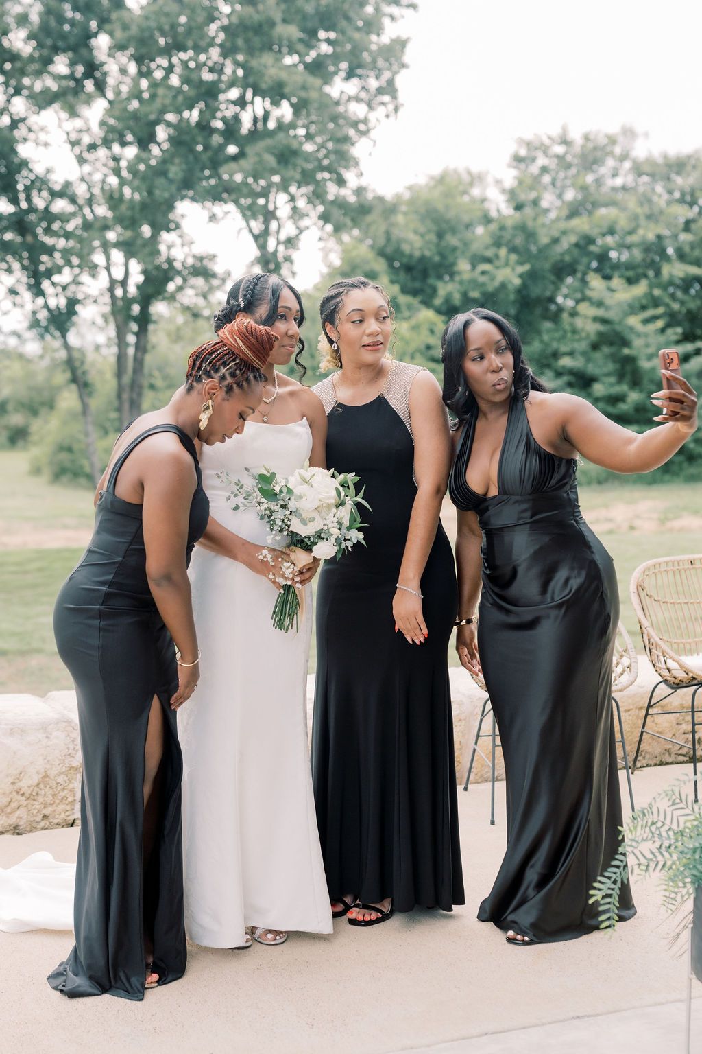 Bride in white gown holds bouquet with three bridesmaids in black dresses taking a selfie outdoors.