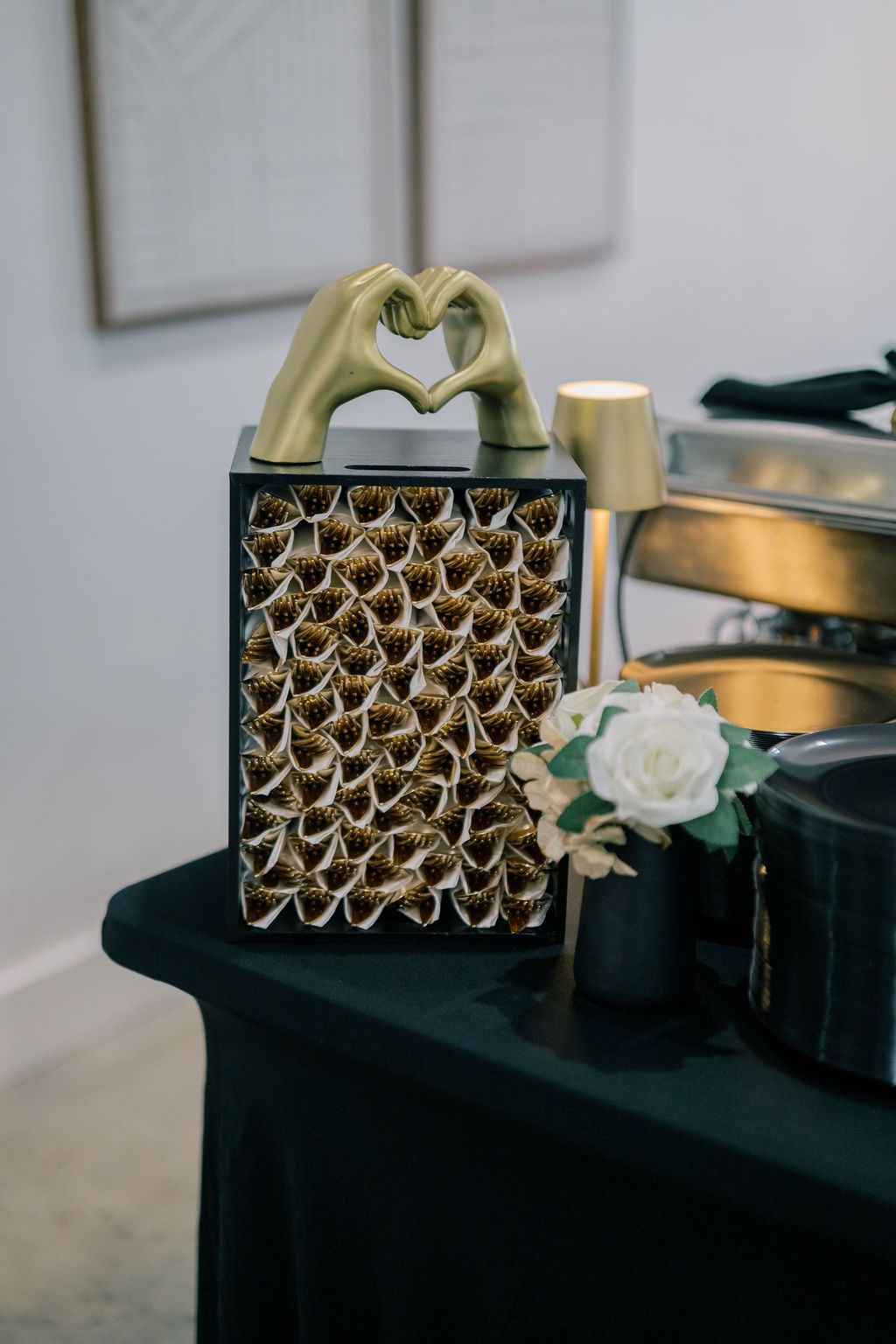 Gold heart hands sculpture atop a black decorative box with floral accents on a dark green draped table.